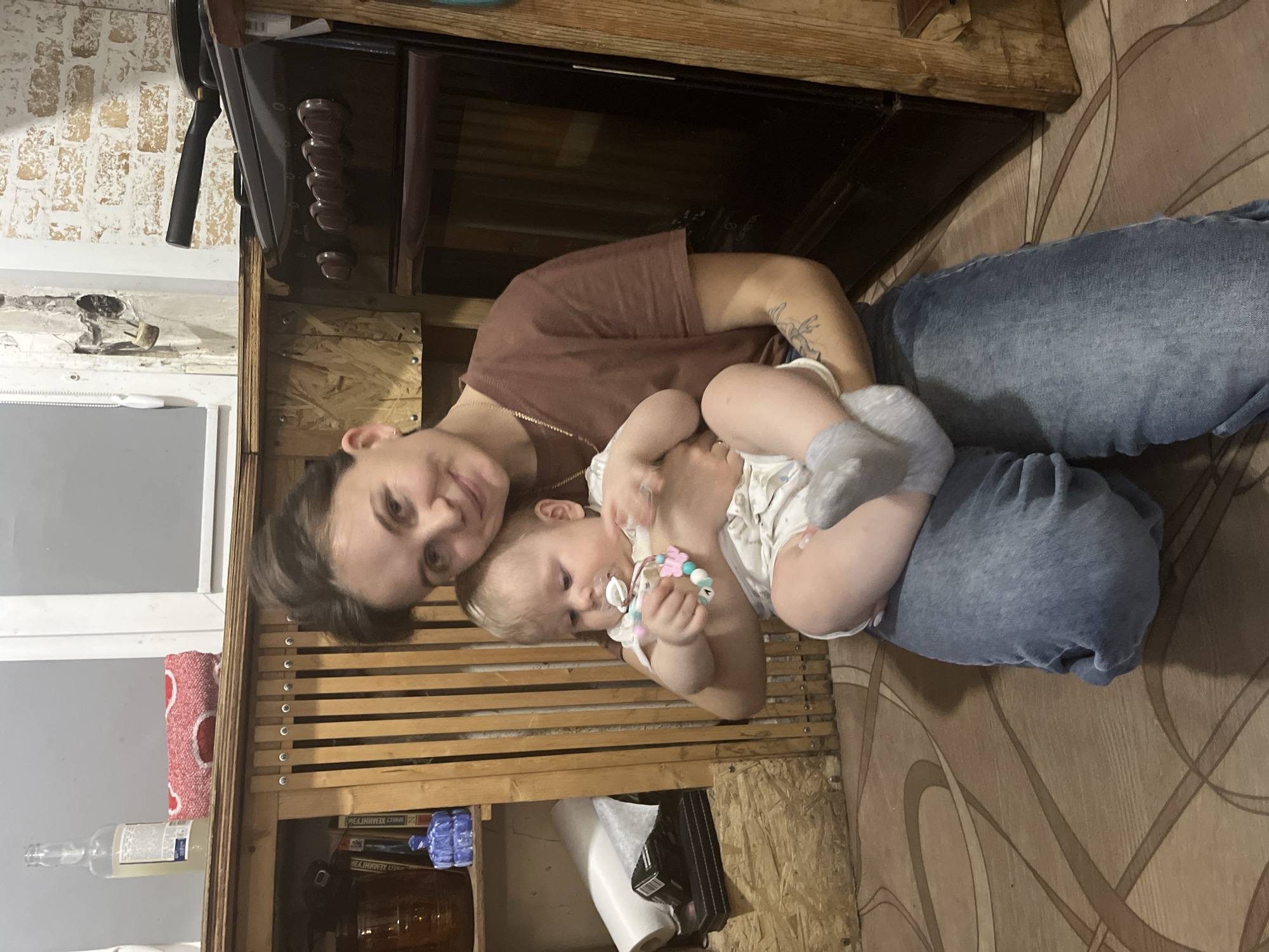 Scene: An interior shot shows a woman holding a baby, both indoors.
People: A woman with dark hair and a brown shirt is holding a baby. The baby is wearing a white and patterned garment with grey socks. The baby holds a pacifier and teething ring.
Objects: The woman wears a gold necklace. The baby has a colorful teething ring and a pacifier in its mouth. Behind them, there is a kitchen oven. A wooden slatted structure is visible, with various items stored inside, including bottles and other containers.
Actions: The woman is holding the baby. The baby is holding a teething ring and pacifier.
Interactions: The woman is holding the baby close.
Environment: The location appears to be a home, specifically the kitchen. The floor has a patterned design. Based on the context of the query, the location is potentially in or around Oryol-Izumrud, Russia, although this is impossible to verify from the image alone.