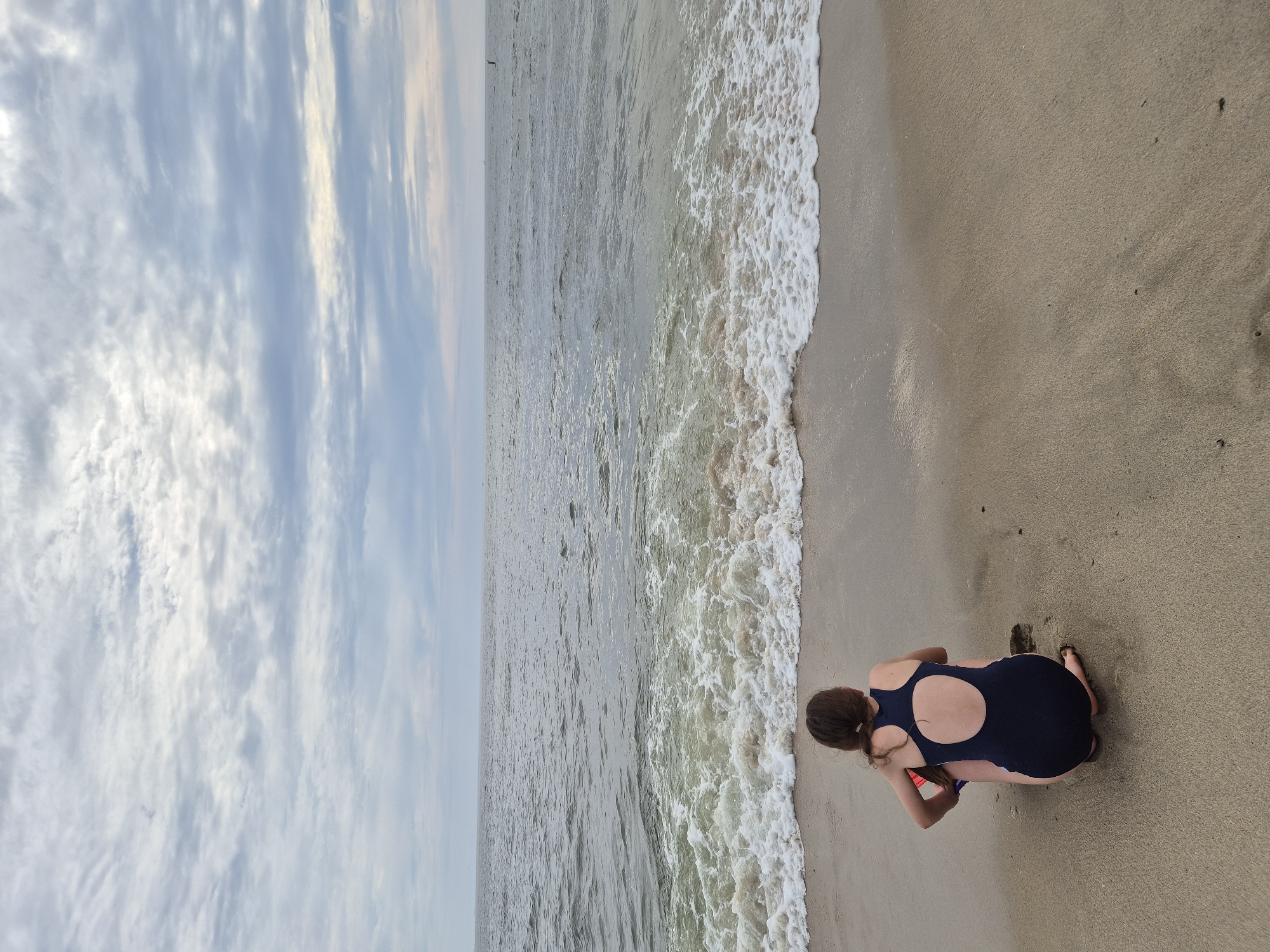 This media file depicts a serene beach scene. The primary subject is a young person wearing a dark blue swimsuit, kneeling on the sandy shore. Their back is to the camera, and their brown hair is pulled back, with a few strands loose around their face. They appear to be digging or playing in the sand, with one hand near the sand and the other tucked underneath. The ocean stretches out before them, with gentle waves breaking on the shore, leaving a foamy white edge along the sand. The water is a muted gray-blue, reflecting the overcast sky.
The sky above is filled with dramatic, wispy clouds, suggesting a cloudy or possibly twilight time of day. The light is soft and diffused, casting no harsh shadows. The sand is a light brown, with some small dark specks and footprints visible. The overall impression is one of quiet contemplation or playful engagement with nature. There are no visible objects other than the person and the natural elements. The location is identified as Middletown Township, USA. The scene conveys a sense of peace and a connection with the coastal environment. The time of day appears to be either early morning or late afternoon, given the soft lighting and cloud cover. The weather seems calm, with a gentle breeze likely contributing to the soft waves.