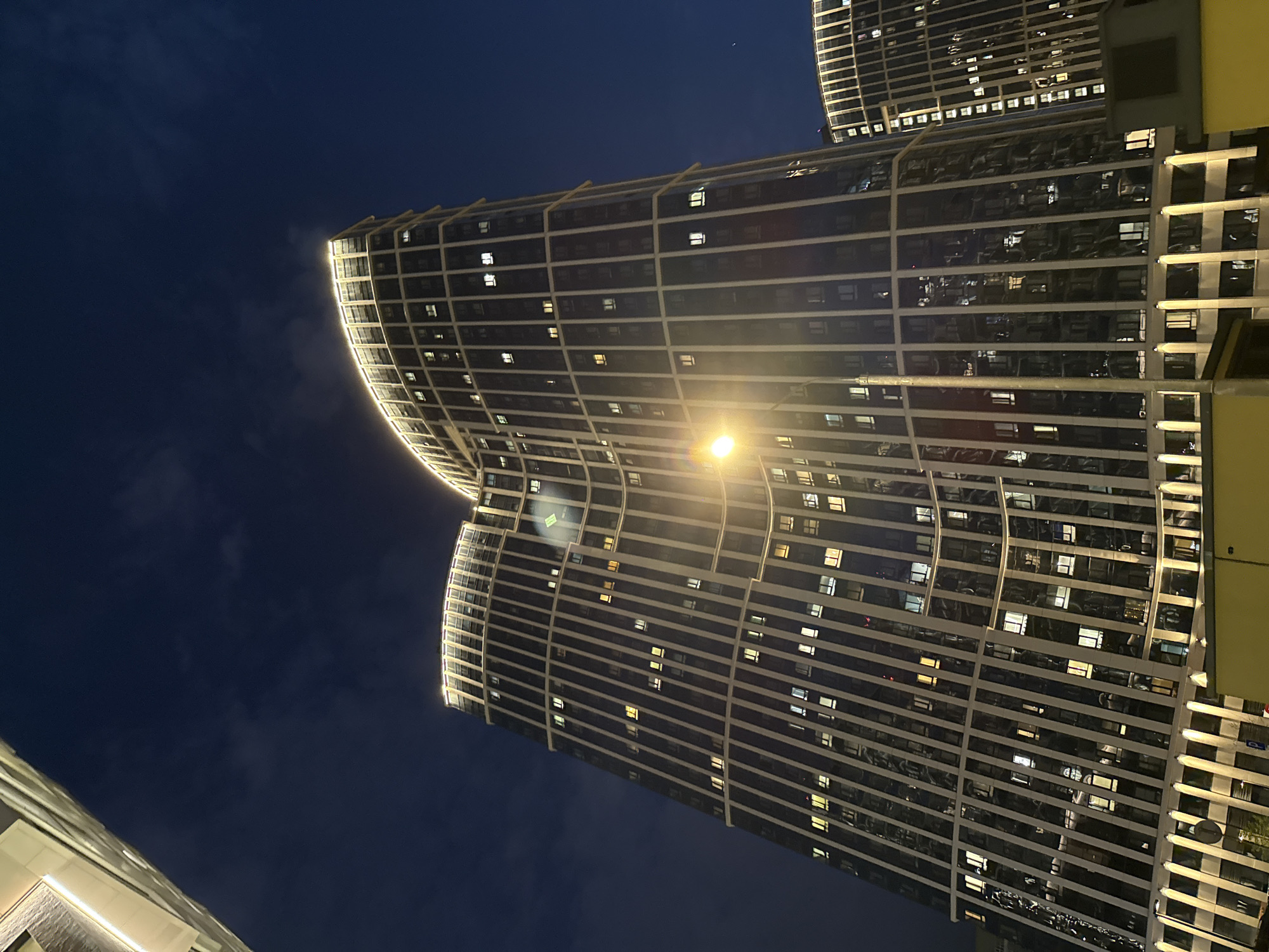 The image depicts a night scene in what is likely Khimki, Russia, featuring modern high-rise buildings with curved facades. The structures are illuminated with interior lights visible through numerous windows. External architectural lighting accentuates the curves of the buildings. The sky is dark, suggesting late evening or night. No people are visible. A light source is present in the foreground, adding brightness to the scene.