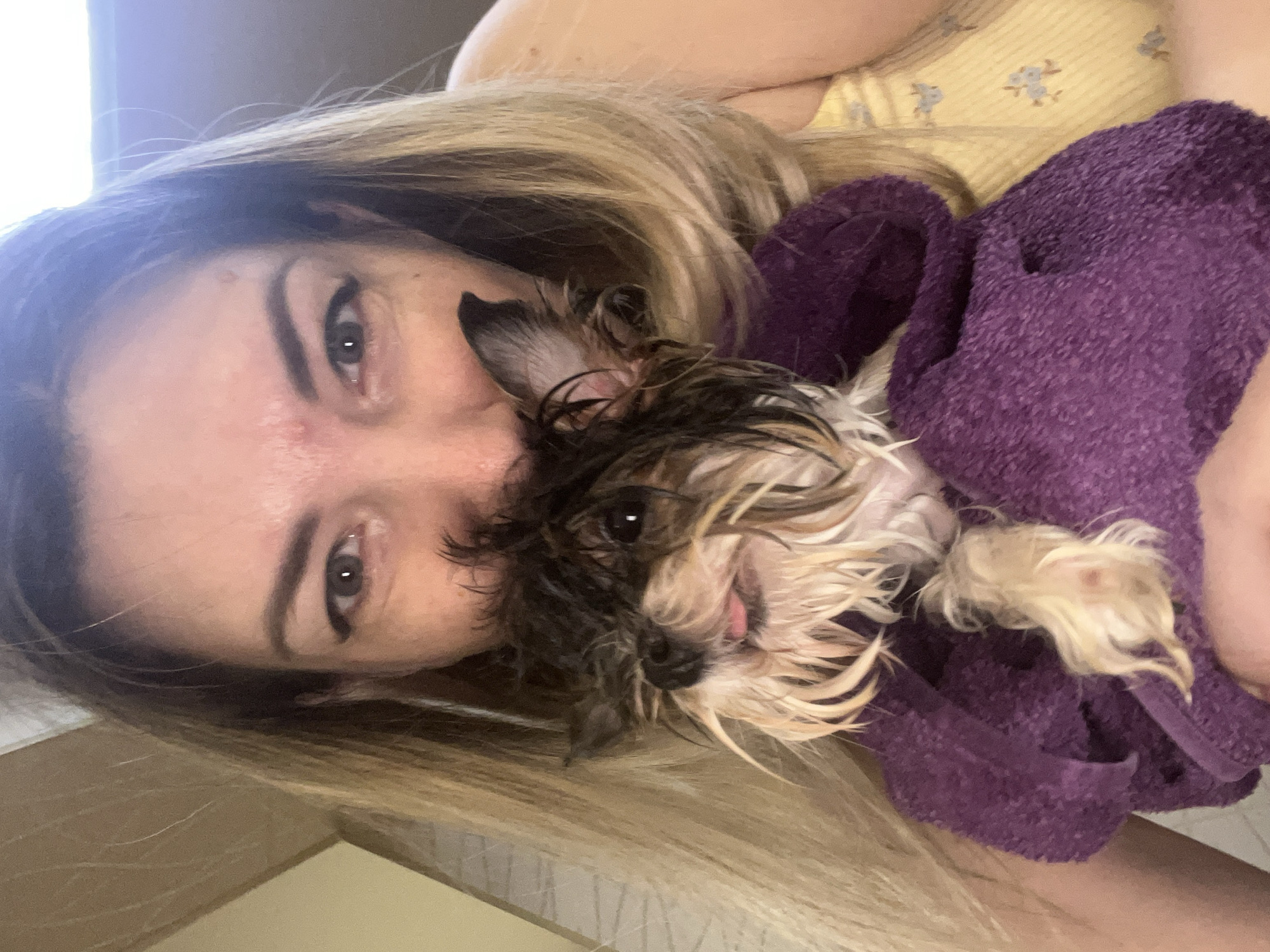 A woman with blonde hair and noticeable makeup is holding a small, wet dog, wrapped in a purple towel. The dog has dark and light fur and its tongue is sticking out. The woman is looking at the camera, and the dog is peeking out from behind her face. The background is indistinct, but appears to be an indoor setting with a wall and possibly a window. The image is a selfie, suggesting the photo was taken by the woman herself. The location is specified as Sochi, Russia, but there are no visual cues in the image to confirm this.