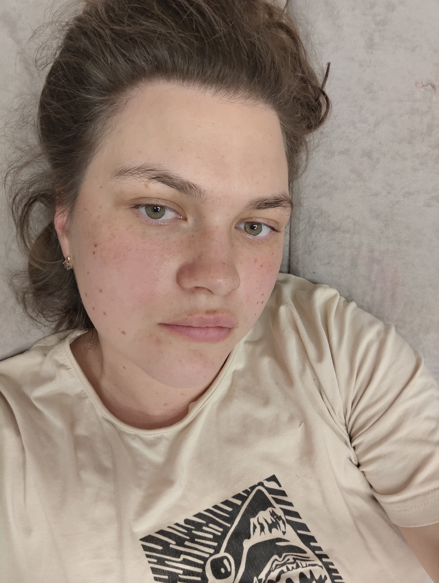 A light-skinned woman with brown hair pulled back in a loose bun is lying on a light gray surface. She has green eyes and freckles on her face. She is wearing a beige t-shirt with a black graphic design on the front. The graphic appears to be a mountain scene inside a geometric shape. There is no discernible information that points to the city of Chaikovsky, Russia.