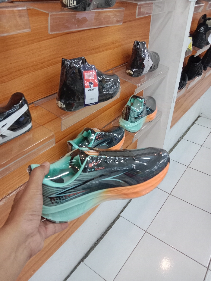 The image depicts an indoor retail environment, likely a shoe store, located in Langenharjo, Indonesia. A human hand, appearing to be an adult's right hand with a medium skin tone, is prominently shown in the foreground, holding a pair of athletic shoes. The shoes being held are black with a light blue/teal and orange gradient sole, wrapped in clear plastic, and bear a visible label on the tongue stating "MADE IN CADILA."
In the background and mid-ground, multiple other pairs of shoes are displayed on clear acrylic shelves mounted onto a light brown wooden slat wall. Several pairs of black, high-top "Ventela" brand shoes, each individually wrapped in clear plastic with product tags, are visible on the upper shelves. Below these, another pair of athletic shoes, similar in design and color scheme to the one being held, rests on a lower shelf. A pair of black and white athletic shoes is partially visible on a shelf to the far left. The floor is composed of white square tiles with dark gray grout lines. The scene is brightly illuminated, with reflections visible on the plastic-wrapped shoes and the tiled floor. No other individuals are visible in the frame.