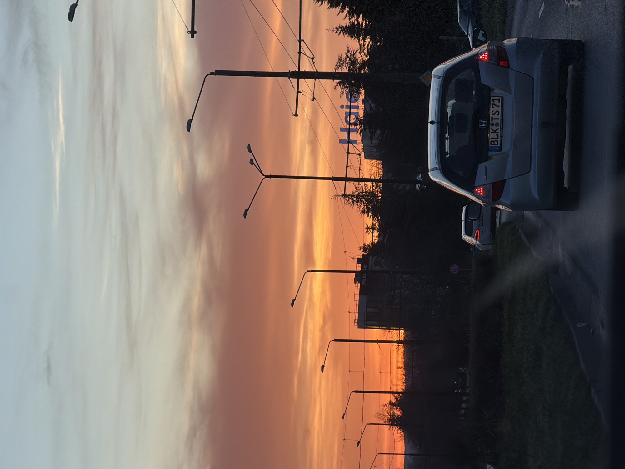 The image depicts a sunset scene in Varna, Bulgaria, viewed from the perspective of someone in a vehicle. A silver Honda Jazz (Fit) with license plate "BLK TS 71" is in the foreground. Streetlights line a road with green vegetation on one side. In the background, the sky is a gradient of orange and yellow due to the sunset, with a hotel sign, possibly "Haier," partially visible against the darkening sky. Overhead electrical lines are also present. The photograph appears to be taken during twilight hours.
