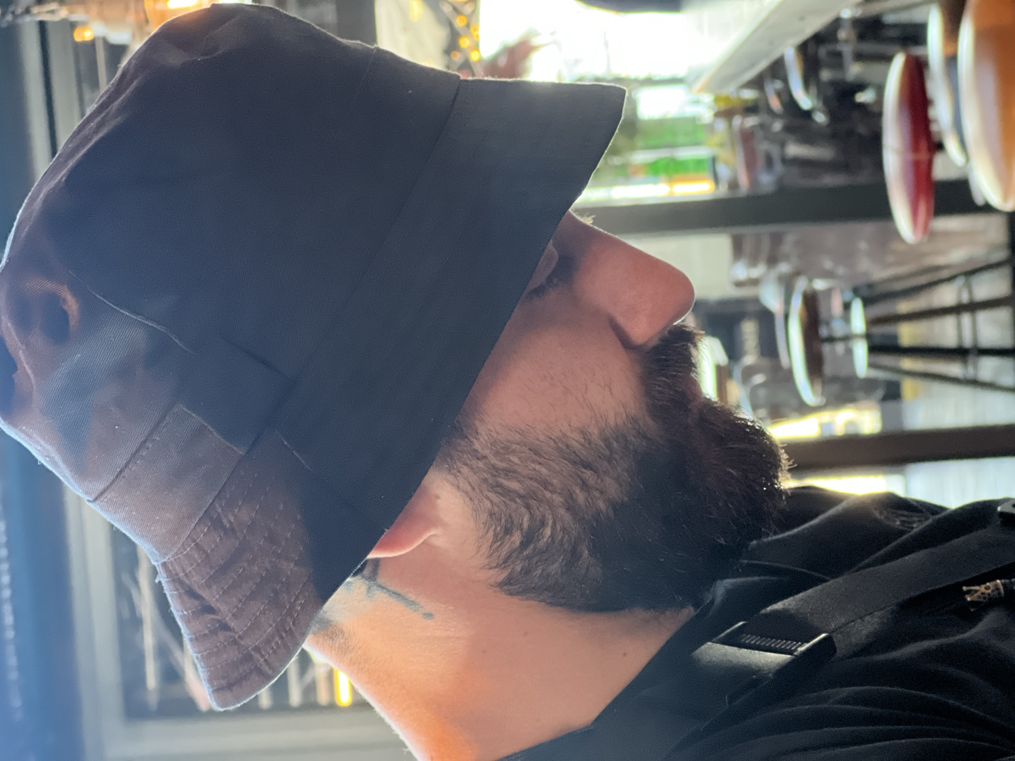 The image is a close-up, angled shot of a person wearing a dark bucket hat and a black shirt, in what appears to be an indoor setting with blurred background elements suggesting a bar or cafe. The person has a beard and a tattoo on their neck. The lighting is warm, with the sun potentially filtering in from the right, casting a glow on the subject's face and neck. The background shows bar stools and what could be bottles on a shelf, implying a public establishment. There is no information about the location in the city of Varna, Bulgaria.