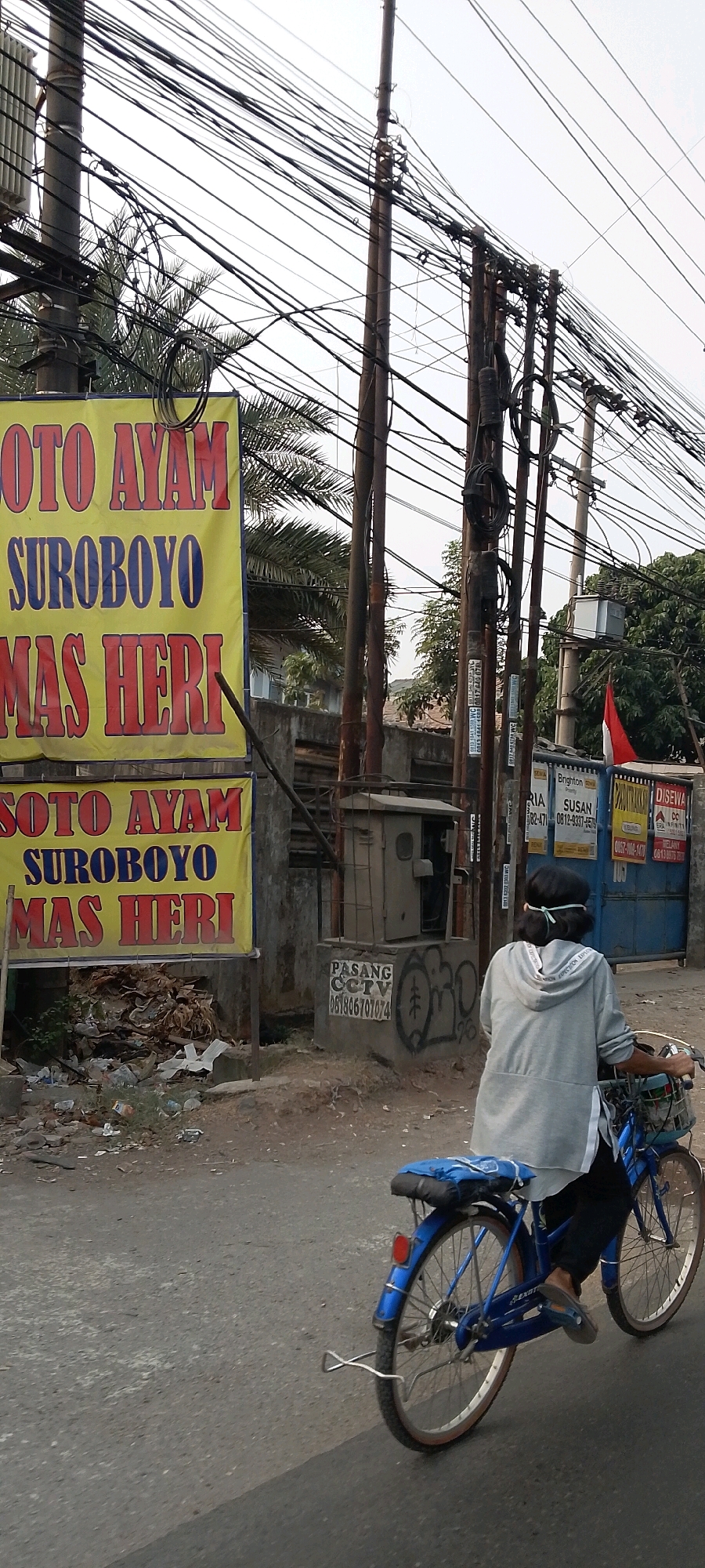 In a cluttered urban scene in Bekasi, Indonesia, a person rides a blue bicycle down a paved street. The cyclist, wearing a gray hoodie and dark pants, has a light blue surgical mask tied around their head. Their bicycle has a basket on the front and a blue bag secured to the rear rack.
The street is lined with utility poles heavily laden with a dense network of electrical wires. In the background, a large yellow banner with red text reads "SOTO AYAM SUROBOYO MAS HERI," indicating a food stall. Adjacent to the banner is a small concrete structure with a metal door, possibly an electrical box, and a sign below it reads "PASANG CCTV" with a phone number. Further to the right, a blue gate is visible with several advertisements attached to it, and a small Indonesian flag is affixed to a pole. The ground along the street is littered with debris and trash. The overall environment suggests a busy, somewhat neglected part of the city.