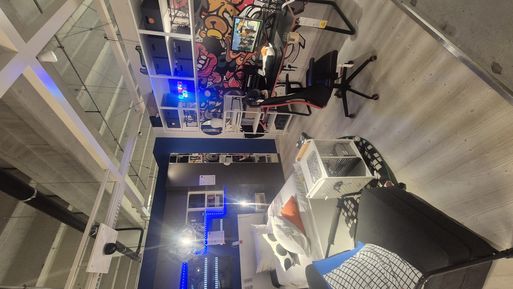 The image depicts a room furnished to showcase a gaming setup and a bedroom. The environment appears to be a retail display, possibly within a furniture store. In the background, a blue wall is visible, with shelves holding various items, including black storage boxes and decorative elements. A neon sign with the words "GAMING ZONE" is prominently displayed on the shelves.
To the right of the neon sign is a gaming desk with a black chair. The desk has a computer monitor displaying a game scene, a keyboard, and other accessories. The chair is a racing-style gaming chair with red accents.
To the left of the gaming setup is a bed with white bedding and several decorative pillows, two of which are white with black polka dots, and one is orange. Adjacent to the bed, a bedside table with a lamp is present. Above the bed, white shelving units are mounted on the wall, illuminated by blue LED strip lights.
In the foreground, a black armchair with a checkered cushion is visible. A white metal trolley cart with shelves containing various items, including boxes and a roll of material, is positioned between the armchair and the gaming desk. The flooring consists of light-colored wooden planks. The ceiling features exposed concrete and industrial-style lighting fixtures and pipes. The overall impression is of a modern, stylized living space designed for both relaxation and gaming. The context of Baronissi, Italy, suggests this might be a display in a local IKEA or similar furniture retailer.