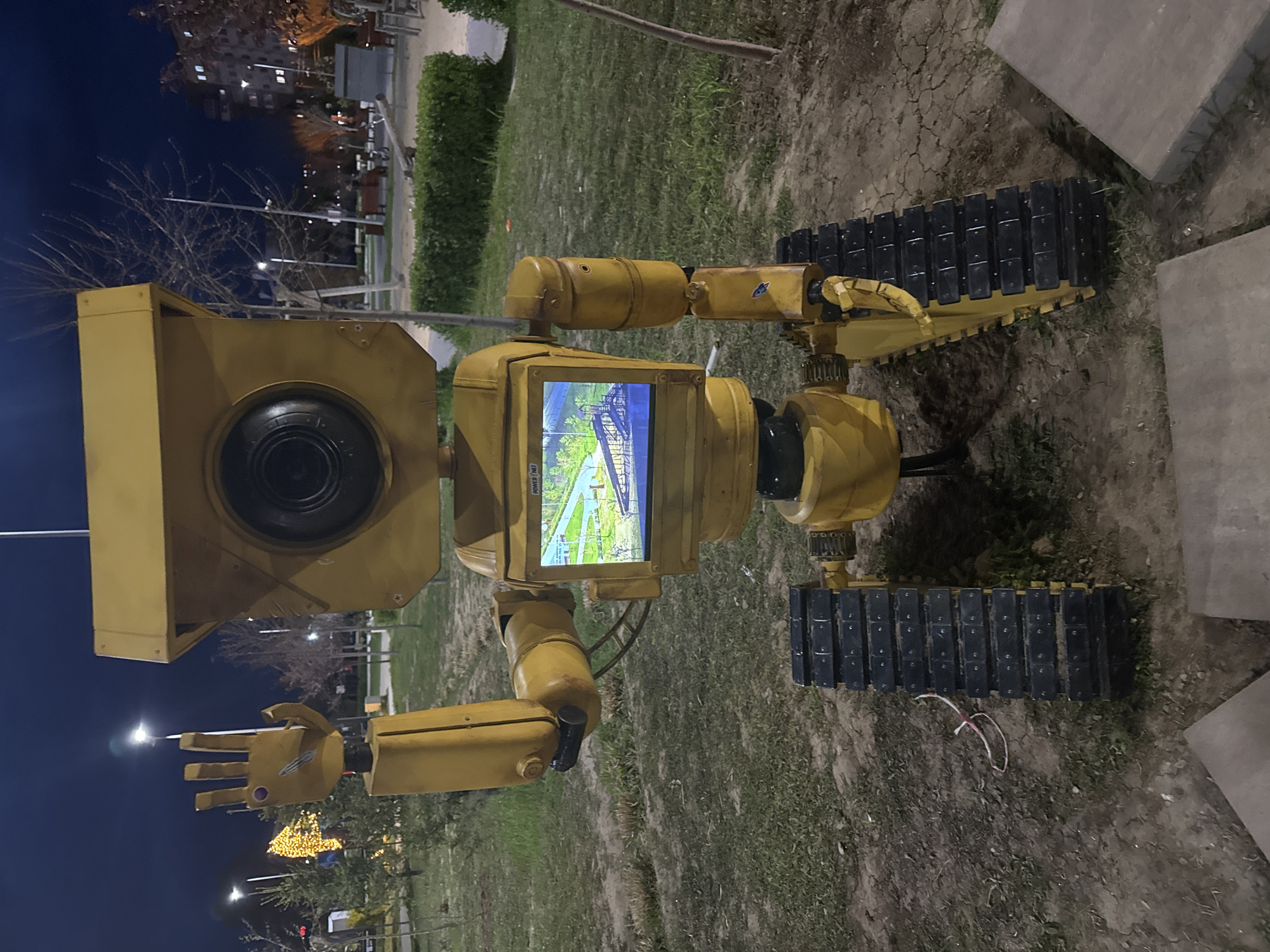 The image depicts a yellow, tracked robot sculpture on a patch of grass, seemingly an outdoor installation, possibly in a park. The robot has a boxy head with a large, dark lens, a screen in its torso displaying a landscape image, and articulated arms with claw-like hands. It rests on two black, tank-like tracks. The background shows a night scene with streetlights, trees, and buildings in the distance. Based on information available online, this likely depicts a robot sculpture located in the city of Volzhsky, Russia. No people are visible in the image.