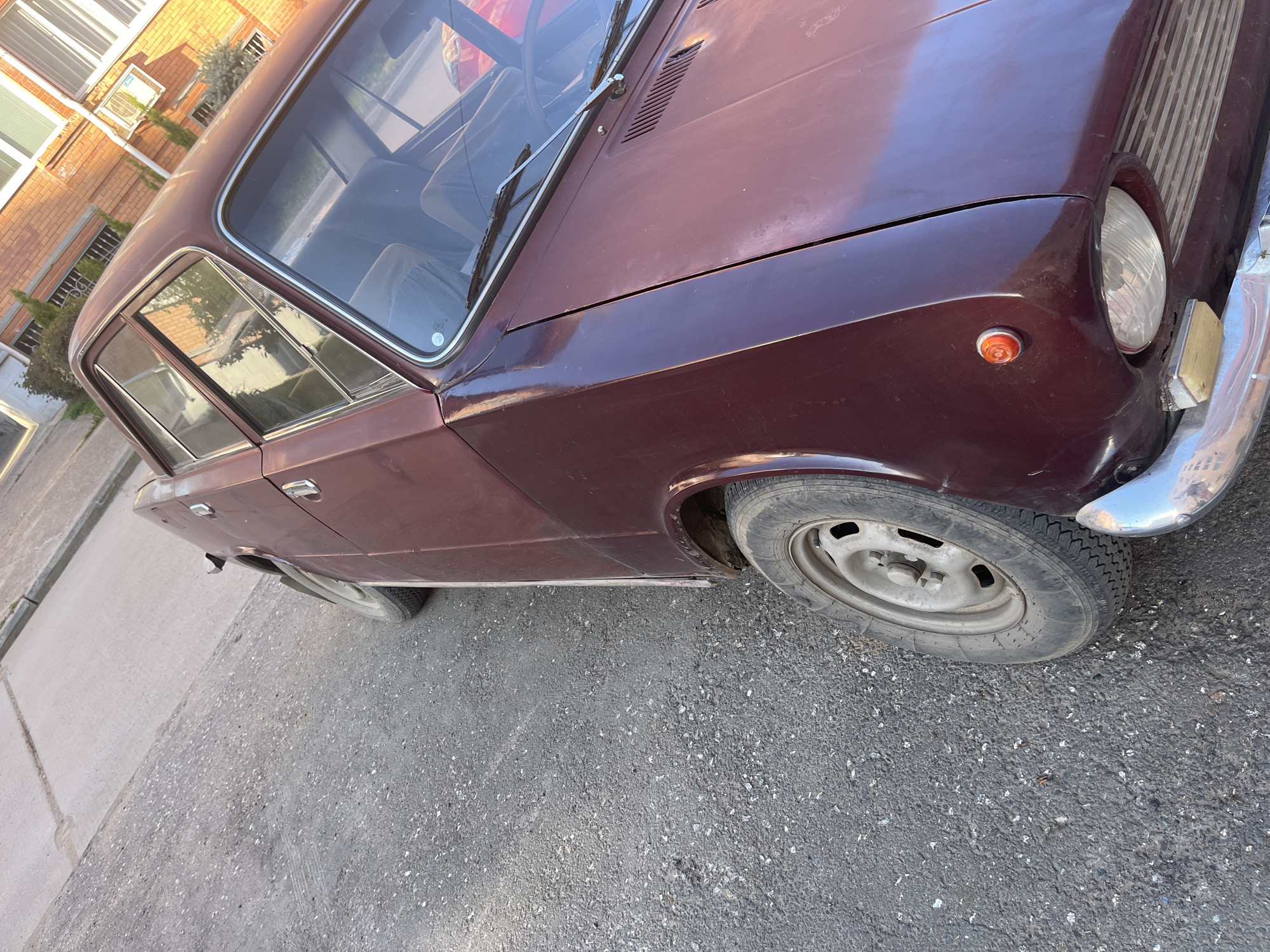 The image depicts a maroon Lada sedan parked on a grey asphalt surface. The car is positioned at an angle, with its front right side facing the viewer. Its tires appear worn, and the body has signs of age and wear. The car's front bumper is chrome. In the background, there's a building with visible windows. The image has a slightly desaturated color palette and is shot during daytime. The location appears to be an urban setting.