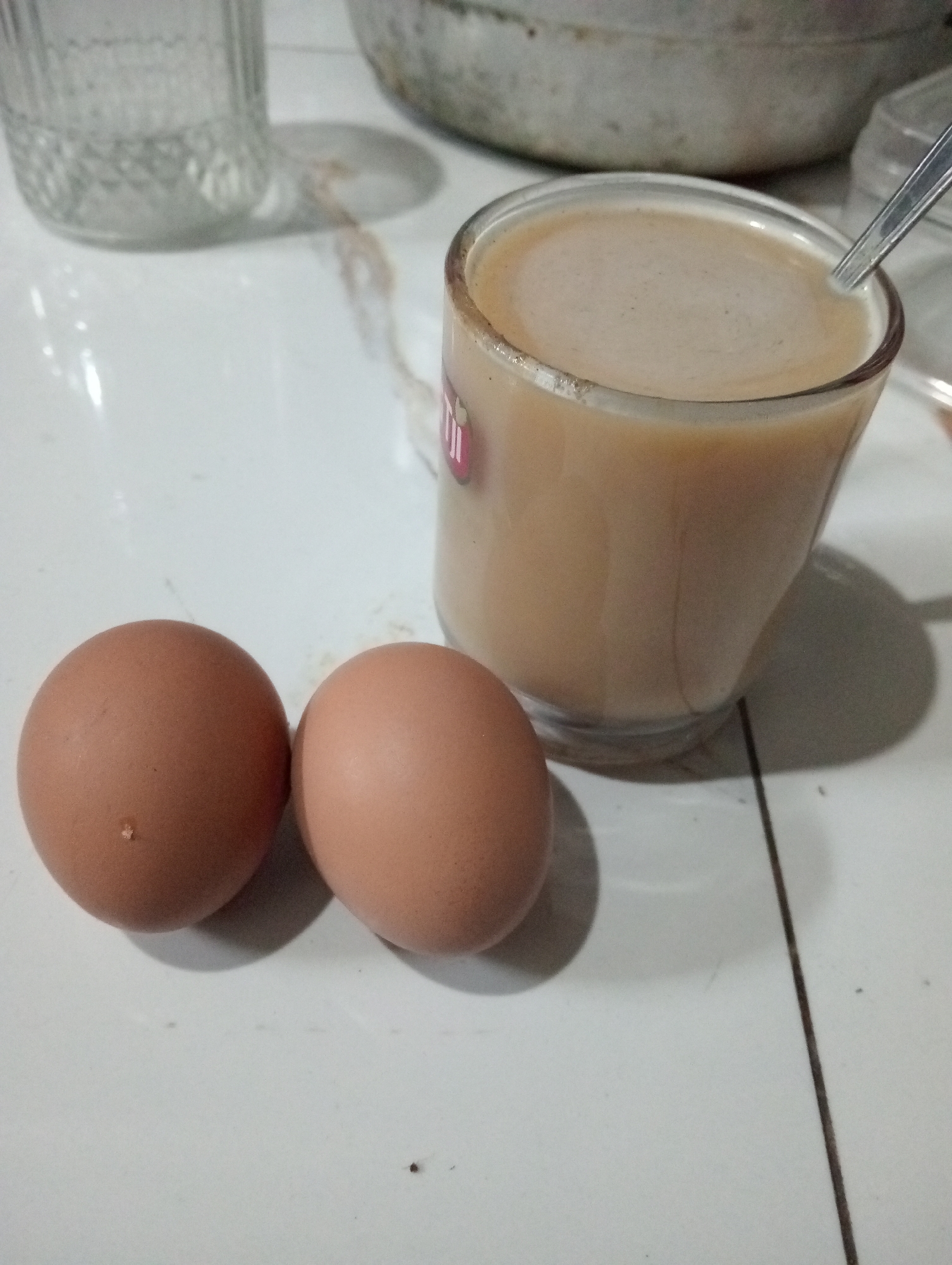 Two brown eggs and a glass of coffee are placed on a tiled surface. The eggs are positioned on the left side of the image, with the glass of coffee to their right. A spoon is partially submerged in the coffee. In the background, a glass bottle and a metal pot are visible. The scene appears to be indoors, likely a kitchen or dining area. The lighting suggests it is daytime. The image does not provide any specific indicators to pinpoint its location within the city of Demak, Indonesia.