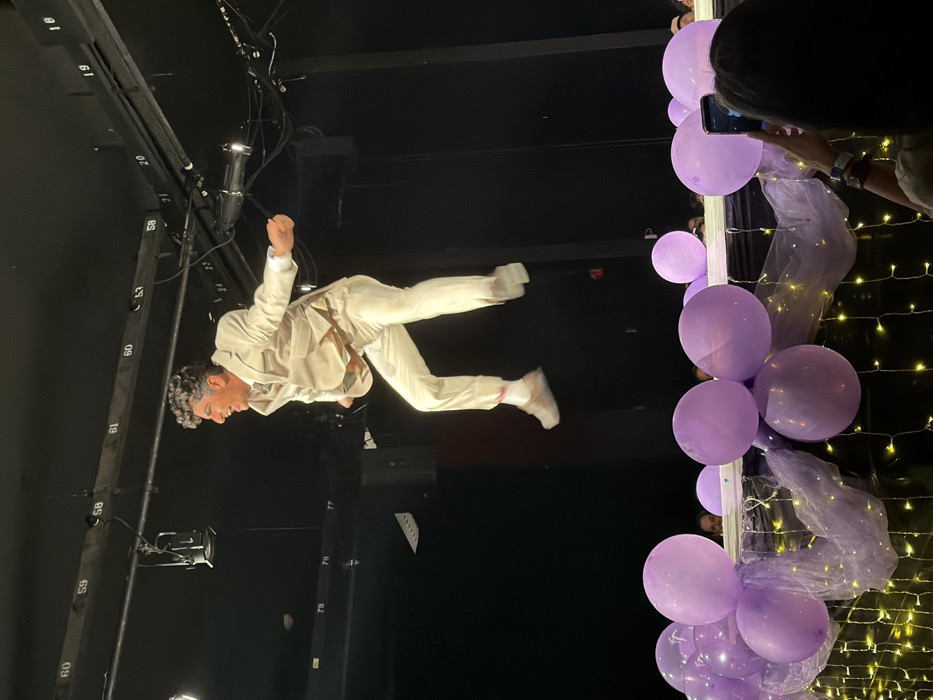 The image captures an indoor stage performance in Palos Verdes Estates, United States. A male individual, appearing mid-air, is the central subject, dressed in a light-colored suit with a light green tie, white socks, and light-colored athletic shoes. His dark, curly hair is visible, and his mouth is open in an expression of excitement or exertion, with both hands clenched. Overhead, exposed metal rigging with stage lights and numerical labels, such as "58" and "59", is visible against a dark background. The lower portion of the frame features a decorated barrier adorned with multiple light purple balloons and sheer purple fabric, illuminated by yellow string lights. Partially visible audience members are situated behind this barrier. In the bottom right corner, a person with dark hair, viewed from behind, is holding a smartphone horizontally, appearing to record the performance.