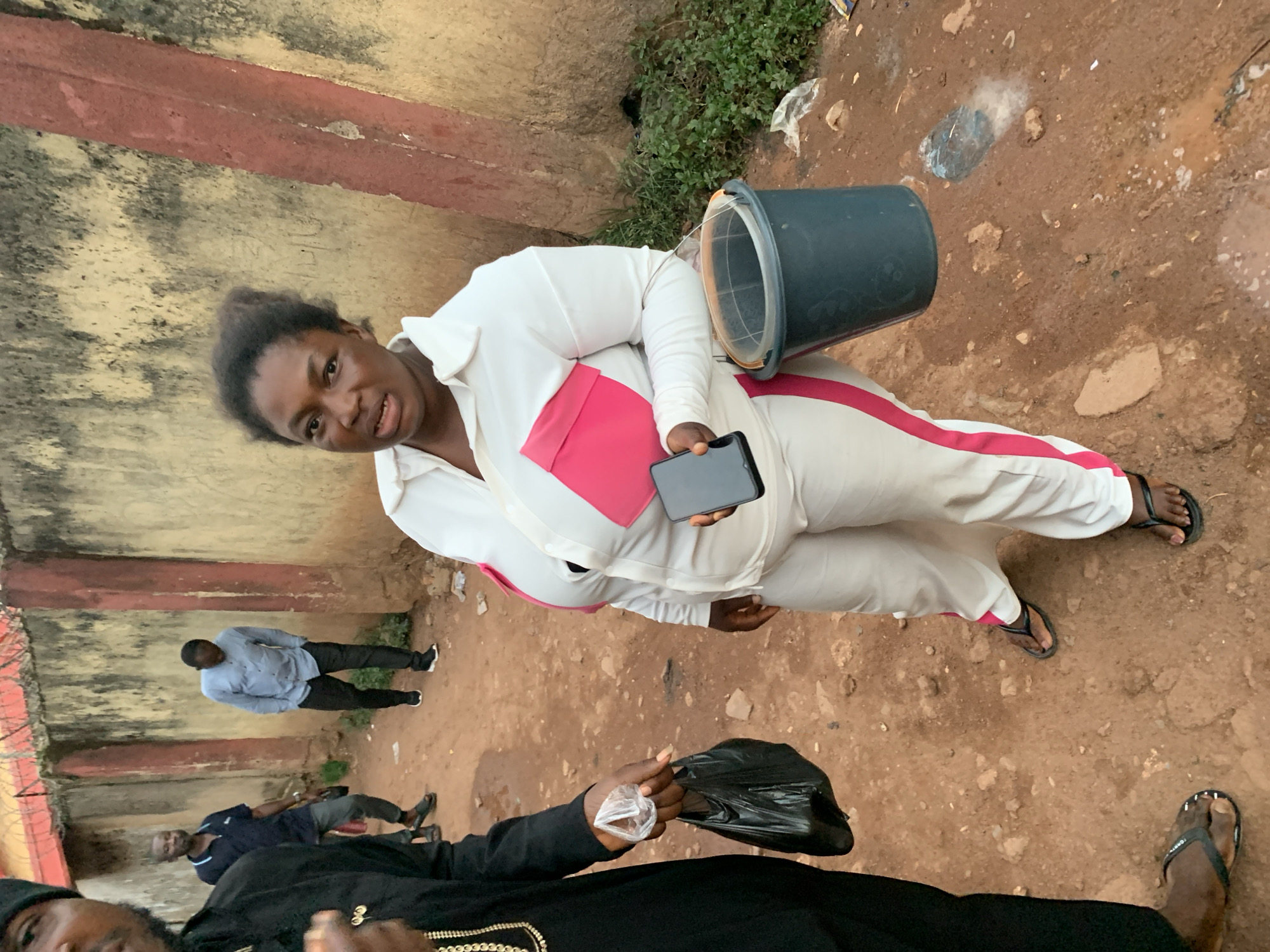 The image shows a group of people outdoors in what appears to be a somewhat neglected urban or semi-urban environment in Jos, Nigeria. The ground is dirt with scattered debris. In the foreground, a person in a black outfit is holding a plastic bag. Behind them, a woman in a white tracksuit with pink accents is holding a dark-colored bucket and a mobile phone. Her expression suggests she is speaking or reacting to something. Further in the background, two men are standing, one in a light blue shirt and dark pants, and another in a dark blue shirt and gray pants. The wall in the background is weathered and shows signs of age. There is some green foliage visible to the right. The scene appears to be taking place during the daytime, and the lighting suggests it could be overcast or late afternoon. The overall impression is of a casual, informal gathering or interaction.