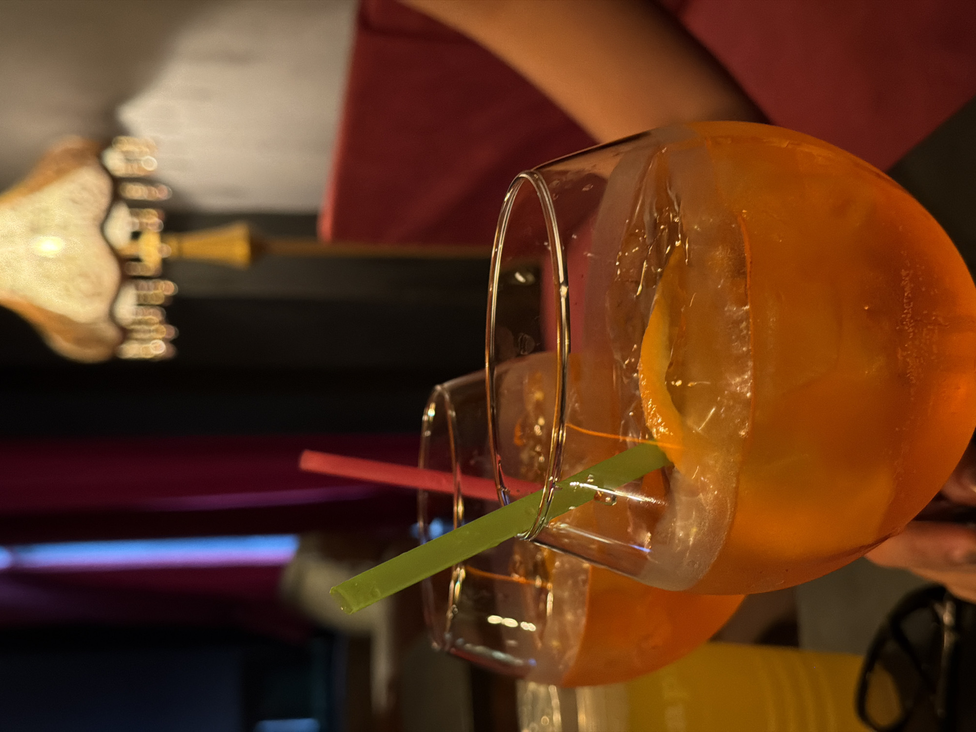 The image shows two glasses of orange-colored cocktails with ice and orange slices. Each glass has a straw, one green and one pink. The glasses are on a dark surface, likely a table. In the background, there is a red curtain, a vintage-style lamp with a yellow base and a lampshade adorned with decorative trim, and a person's arm, suggesting the cocktails are being held or presented. There's also a yellow bottle or container partially visible on the left. The overall setting appears to be indoors, possibly a bar or restaurant, with subdued lighting. The image does not provide any specific information that would allow for identification of the location as Bishkek City, Kyrgyzstan.