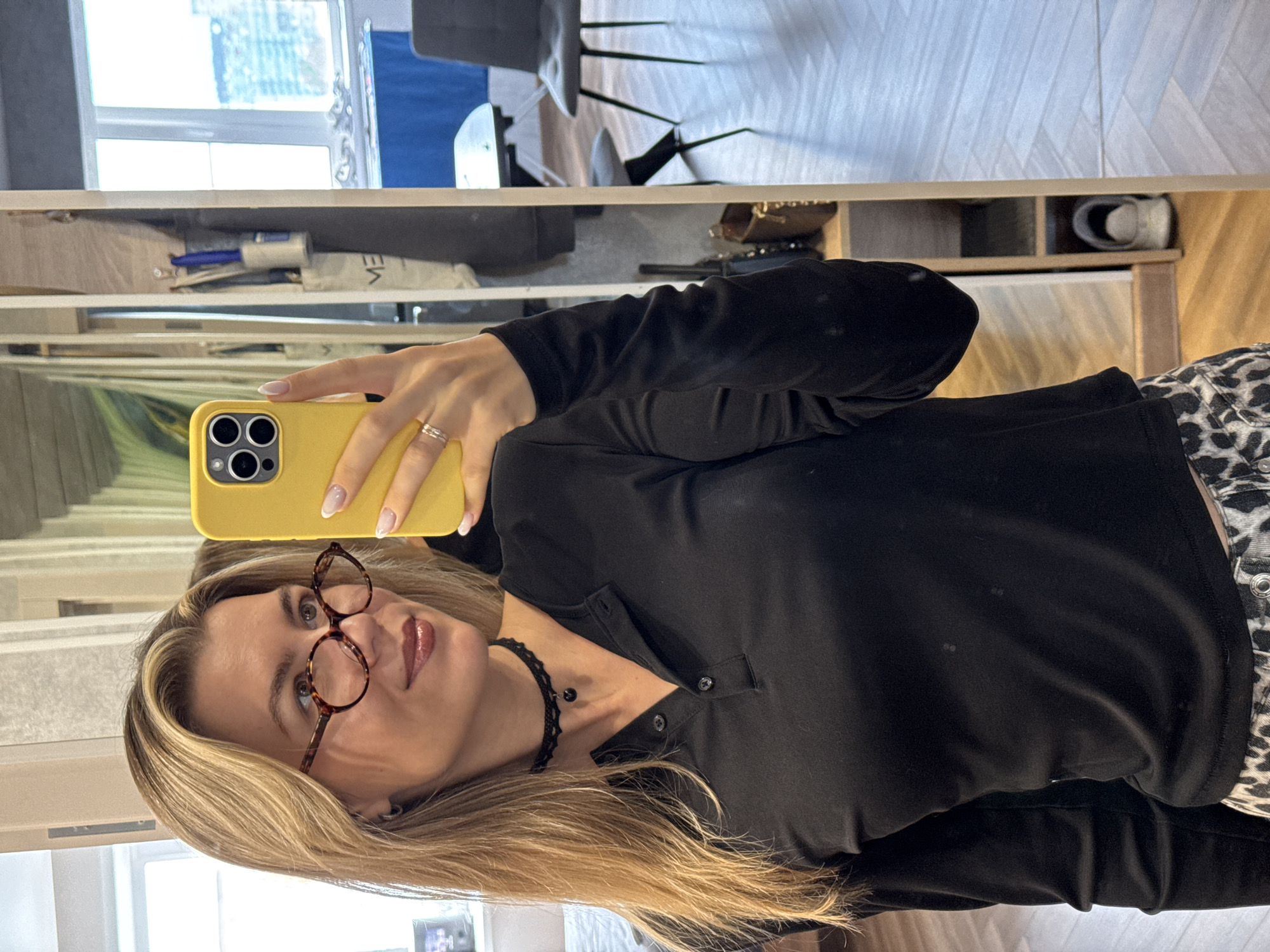 A woman is taking a selfie in what appears to be an apartment or home setting. She is holding a yellow smartphone in her right hand, capturing her reflection in a mirror. The woman has blonde hair and is wearing glasses, a black shirt with buttons, and a black choker necklace. Behind her, the reflection shows various items on shelves, including storage boxes, a lint roller, and other miscellaneous objects. There's also a glimpse of a room with a chair and a window in the background. The city and specific location are unknown.