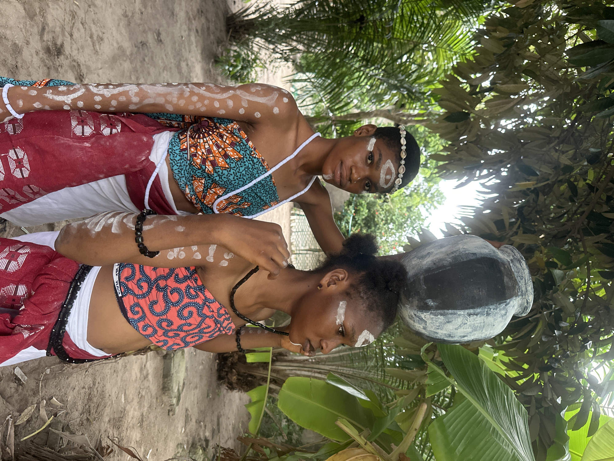 In an outdoor setting, possibly in Badagry, Nigeria, two young individuals are depicted. The person on the left, facing the camera, has their hair styled with a seashell headband and white markings on their face and arms. They are wearing a strapless, patterned top in teal and orange hues, with a white cloth and a red sash around their waist. To the right, another individual, seen in profile, also has white markings on their face and arms and wears a similar strapless top, this one in red with a blue pattern. This person is holding a dark necklace. The person on the left is also holding a large, dark, textured pot on their head, and their right hand is gently touching the head of the person on the right, possibly adjusting the pot. The background features lush green foliage, including palm leaves and other trees, suggesting a tropical environment. The ground appears to be sandy or dirt. The lighting indicates it is daytime.