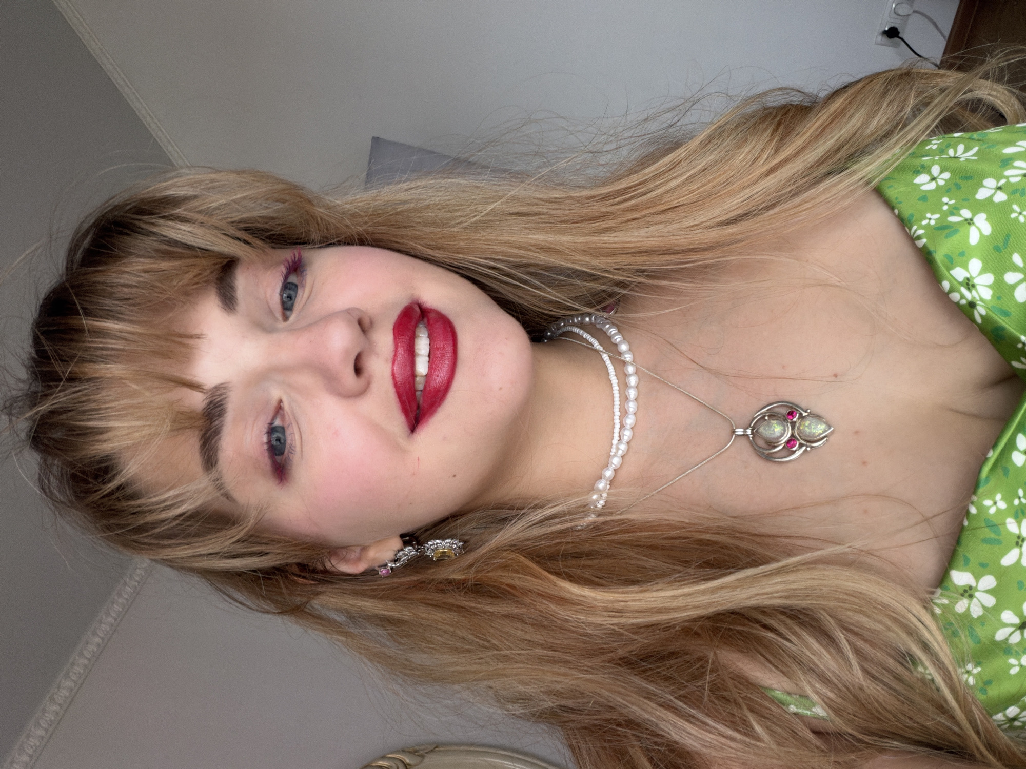 A medium shot depicts a fair-skinned young woman with blonde hair and blue eyes. She is wearing a green dress with a white floral pattern and jewelry including a pearl necklace, a pendant, and earrings. She is wearing makeup including red lipstick and pink eyeshadow. The location appears to be indoors, with a wall and electrical outlet visible in the background, potentially within the city of Большая Шильна, Russia. The image is rotated counterclockwise.