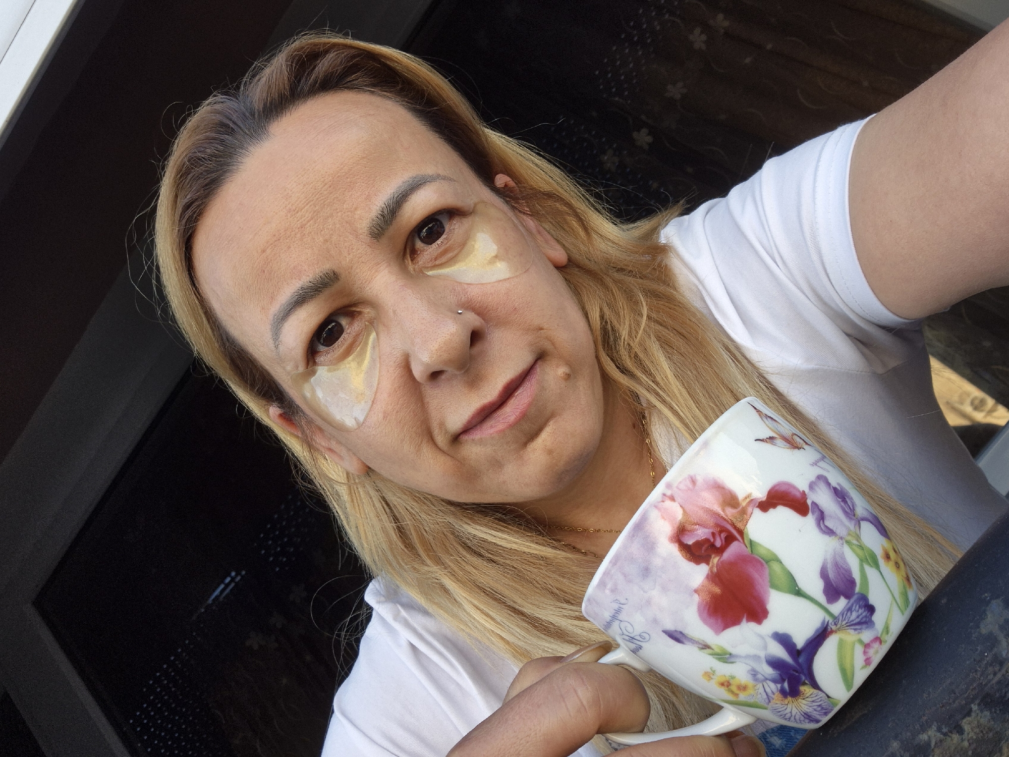 A woman with blonde hair and under-eye patches holds a floral-patterned mug in her left hand. She is wearing a white t-shirt. The background is dark and indistinct, with hints of a window or screen. The image appears to be a selfie taken outdoors.