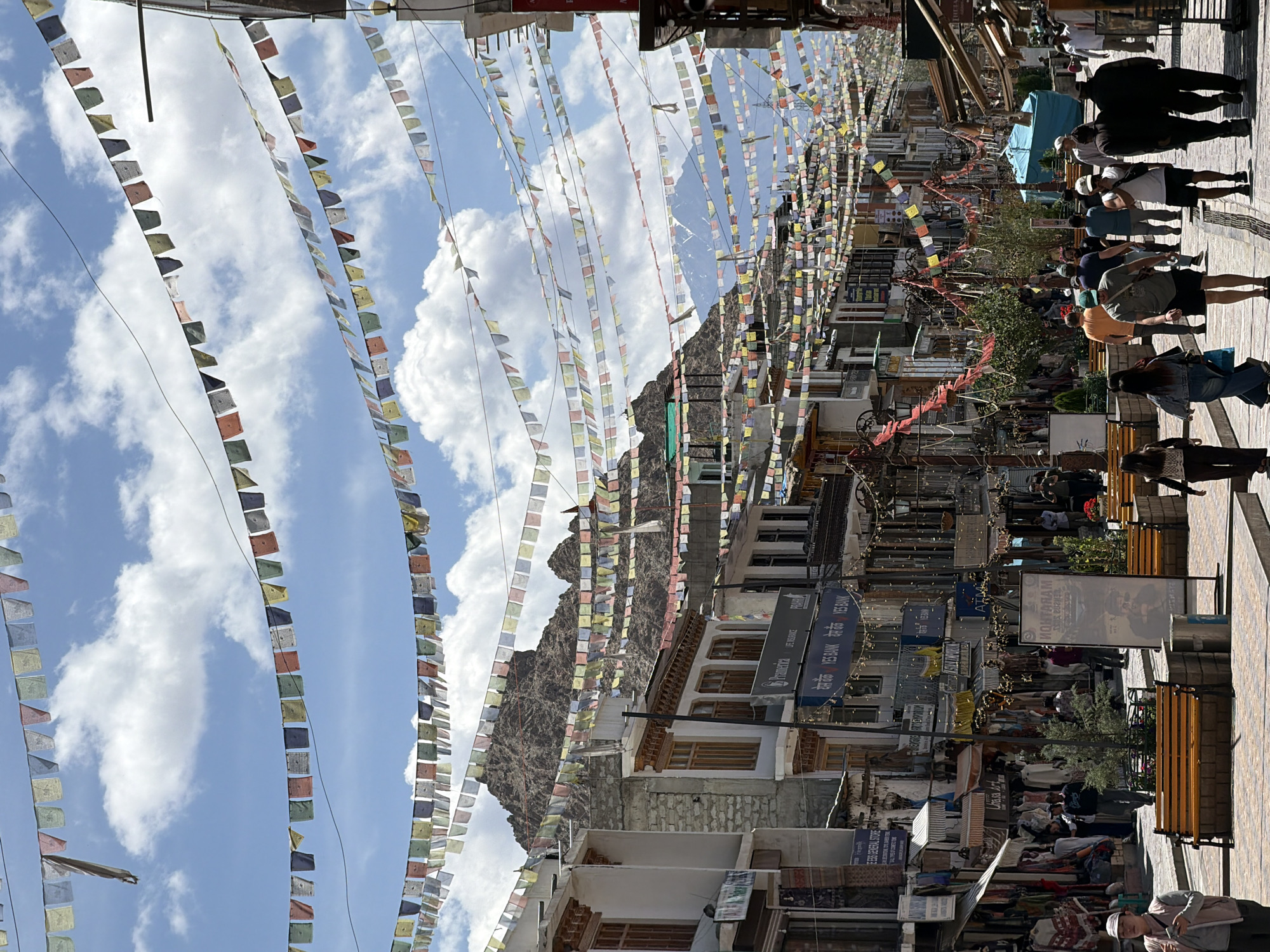 The image depicts a bustling street scene in the city of Leh, India, under a partly cloudy sky. Prayer flags are strung across the street, creating a colorful canopy overhead. The buildings lining the street are a mix of traditional and modern architecture, with shops and businesses visible on the ground floor. People are walking along the street, shopping, and interacting with each other. The scene suggests a vibrant and active urban environment. The presence of prayer flags is a strong indicator that this is a location with a significant Tibetan Buddhist influence, common in Leh, which is the capital of the Ladakh region. The rugged mountains in the background further reinforce this geographical context.