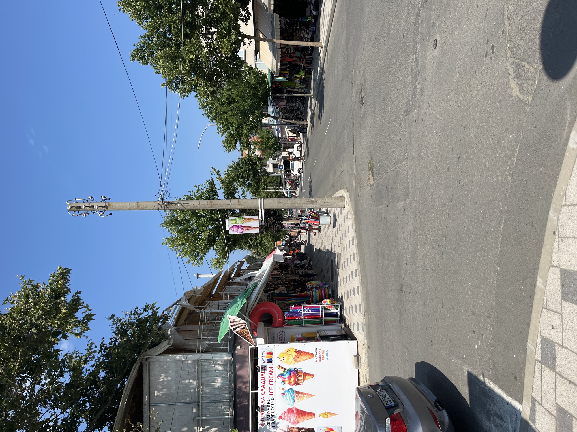 The image depicts a street scene in Obzor, Bulgaria, likely during daytime given the bright sunlight and clear blue sky. The foreground is dominated by a paved road with a gray car parked on the left. To the right of the car, a sidewalk features a patterned tile design. Along the sidewalk, there are several market stalls selling various goods.
One stall prominently displays an advertisement for ice cream with colorful images of cones. Adjacent to this stall, beach inflatable toys and other merchandise are visible. Further down the street, more market stalls are set up, with awnings providing shade. People can be seen browsing the shops and walking along the sidewalk. Trees line the street, offering shade and contributing to the pleasant atmosphere. In the background, a utility pole with electrical wires stands tall. The overall impression is that of a bustling commercial street in a seaside town, likely a tourist area.