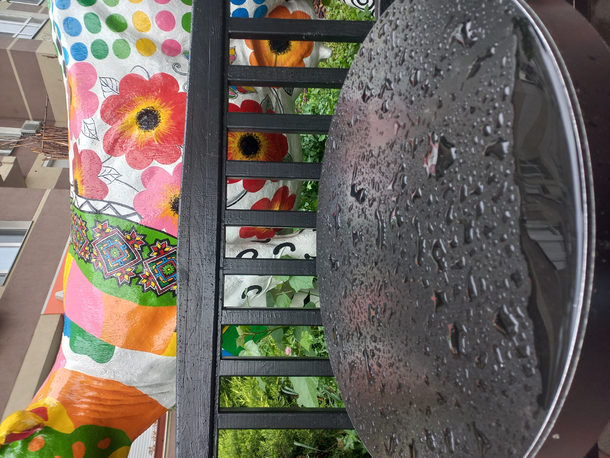The image captures an outdoor scene, likely in Kyiv, Ukraine. The primary focus is a round, dark-colored table covered in raindrops, suggesting a recent rainfall. Behind the table, a black fence partially obscures a brightly colored, cylindrical object adorned with floral and geometric patterns. The object's base appears to be painted with various hues like orange, green, pink, and white. Through the gaps in the fence, green vegetation and glimpses of a building in the background are visible. The composition suggests the image was taken in a public space like a park or sidewalk cafe, capturing the contrast between the functional (table and fence), decorative (painted object), and natural elements (vegetation and raindrops).