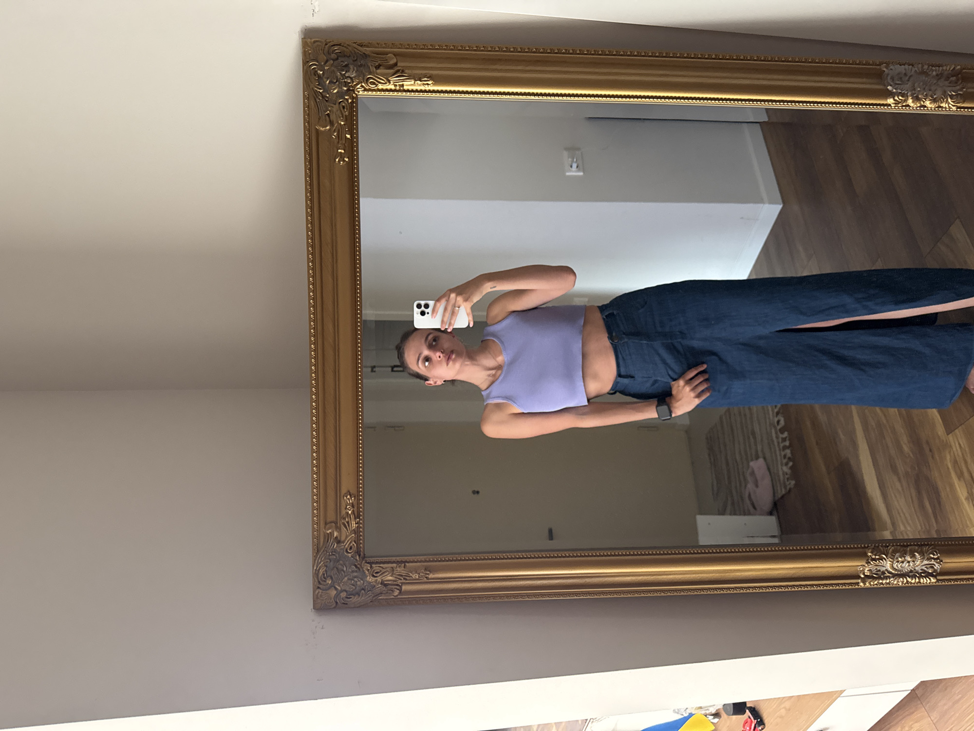 A young woman is captured in a mirror selfie, with the image rotated 90 degrees clockwise. She is light-skinned, has her hair pulled back, and wears a lavender sleeveless crop top and dark, wide-leg jeans. Her left arm is raised, holding a white smartphone with its camera pointed towards the mirror to take the photo. A ring is visible on her left hand, and she wears a dark-banded smartwatch on her left wrist. A small tattoo is visible on her left bicep.
The mirror she is using has a large, ornate gold-colored frame with decorative carvings, particularly prominent at the corners. The reflection in the mirror reveals an indoor setting with light-colored walls and wood-look flooring. An electrical outlet is visible on the wall in the reflection. A light-colored object, possibly clothing or a bag, rests on the floor within the reflection. The area outside the mirror's frame consists of a plain light-colored wall. At the very bottom right corner of the original image, partially cropped, some colorful items like a yellow and blue object and a small red toy are faintly discernible. The woman's expression is neutral. No discernible text is visible.