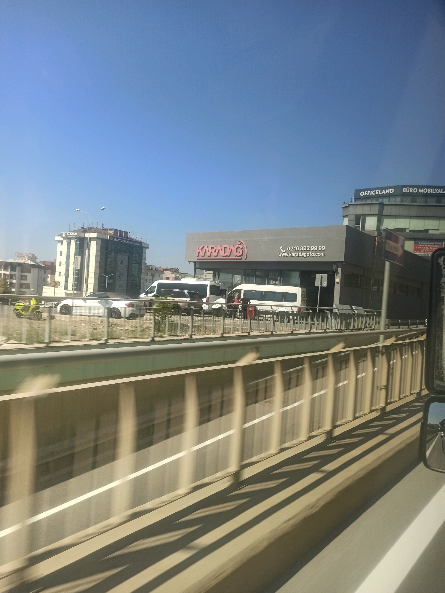 This image depicts a street scene in Pendik, Turkey, captured from a moving vehicle. The foreground is dominated by a concrete barrier and the blurred motion of the road. In the midground, there is a street with several vehicles, including a green scooter on the far left, followed by white cars and vans. A white bus with tinted windows is parked on the right side of the street. A building with a large sign that reads "KARADAĞ" is visible behind the bus, featuring a phone number and website address. To the right of the "KARADAĞ" building, another building with "OFFICELAND" and "BÜRO MOBİLYALARI" signage is present. The sky is clear and blue. The overall scene suggests a commercial area within the city, likely along a main road.