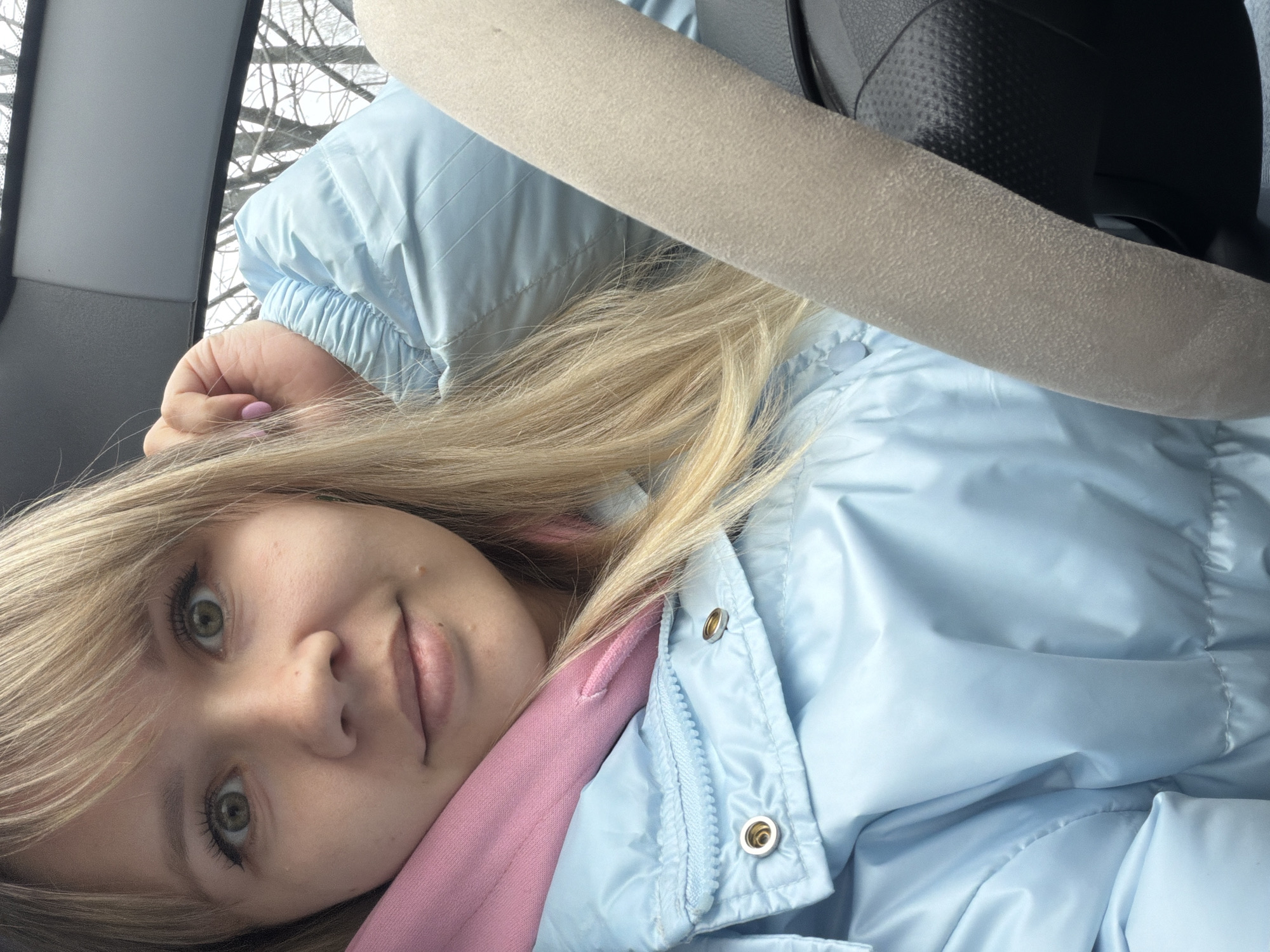 A close-up, angled shot features a young woman inside a vehicle, possibly a car, with the image rotated sideways. She has blonde hair, styled with bangs, and striking green eyes. Her expression is calm, with a slight, subtle smile on her lips. She is wearing a light blue puffer jacket over a pink hoodie.
The background visible through the car window shows bare tree branches against a bright, overcast sky, suggesting a winter or early spring setting. Part of a grey car interior panel is visible to the left. A beige seatbelt crosses the upper right portion of the frame, partially covering her hair and the jacket. Details of the car seat's dark, textured material are visible behind the seatbelt. Her right hand, with light pink nail polish on one visible finger, is resting near her hair. The lighting is natural and soft, likely coming from outside the vehicle during the day. No visible text is present in the image.