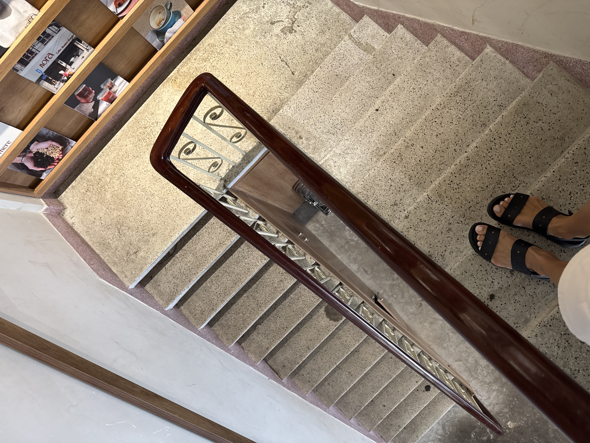 An overhead shot shows a person's feet wearing black sandals, standing on a terrazzo staircase with a dark wooden handrail. The staircase leads down into a building, with shelves displaying various magazines and prints on the right wall. A light-colored wall borders the staircase on the left. The scene appears to be in Nha Trang Ward, Vietnam.