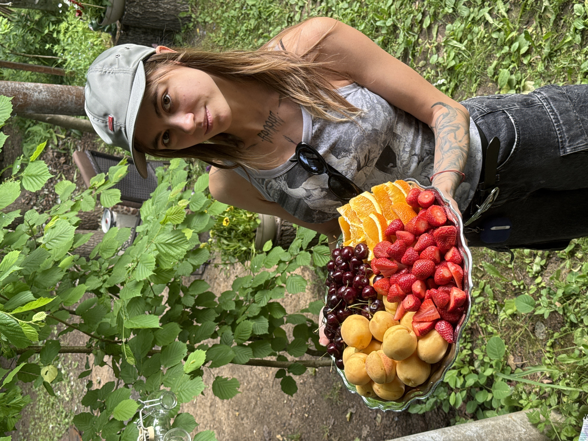 A young woman wearing a gray baseball cap and a gray tank top with a bird print is holding a large silver platter of fruit. The platter contains apricots, cherries, sliced oranges, and strawberries. She has tattoos on her arms and chest. She is standing in an outdoor setting with green trees and foliage in the background. The ground is covered with grass and dirt. The image appears to have been taken during the daytime. The specific location is Буран Снт, Russia.