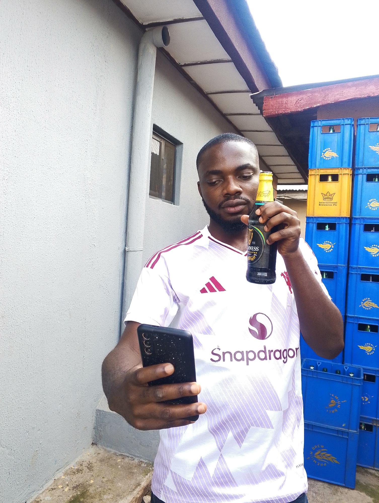 A man stands outdoors, holding a bottle of Guinness in his right hand and a smartphone in his left. He is wearing a white t-shirt with maroon accents and the "Snapdragon" logo. Behind him, stacks of blue and yellow beer crates are visible. The scene appears to be in a somewhat industrial or storage area, possibly a warehouse or a beverage distribution point, given the crates. The lighting suggests it is daytime, and the overall impression is casual, possibly a moment captured during a break or while documenting something with his phone. The background wall is textured and painted light gray, with a downpipe running vertically. The roofline and wooden beams are visible overhead. The man's expression is neutral, focused on his phone and the beer bottle. The text "Guinness Extra Stout" is clearly visible on the bottle, and "Snapdragon" is prominent on his shirt. The crates are marked with text including "BREW" and "PREMIUM". The location context provided places this scene in Jos, Nigeria.