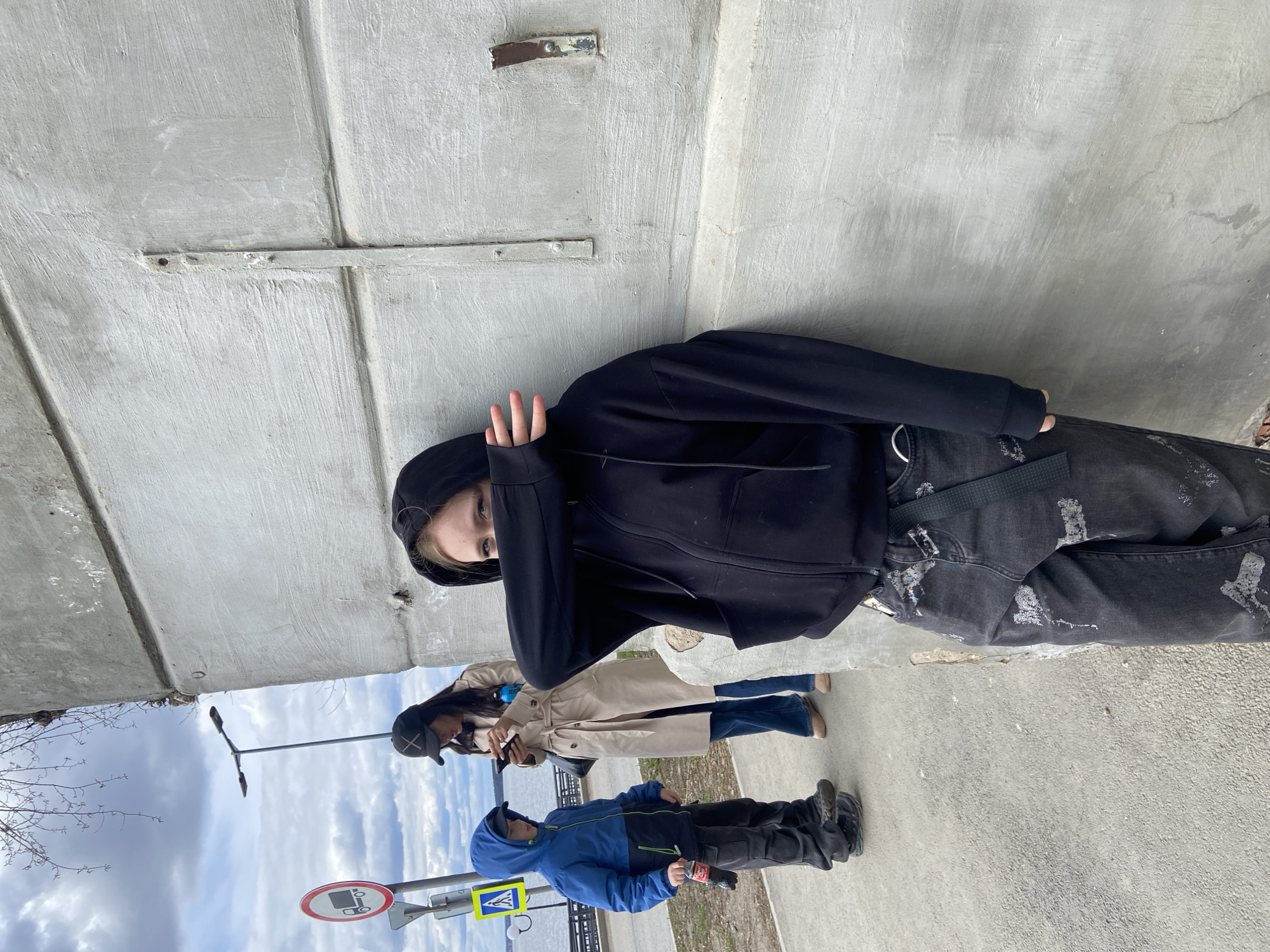 The image captures three individuals standing outdoors next to a large concrete wall or structure, likely a bridge support or an underpass. The scene suggests an urban or semi-urban setting, possibly along an embankment or promenade, with a body of water visible in the background.
In the foreground, a young person wearing a black hooded jacket and distressed dark jeans stands against the concrete wall, partially obscuring their face with a hand, looking towards the viewer. They have a black utility belt with a silver buckle and a black strap across their jeans. Behind them, a woman in a light-colored trench coat, jeans, and a dark baseball cap with a prominent 'X' is visible. She holds a smartphone and has a blue bottle attached to her bag. To her left, a child in a blue and black hooded jacket and dark pants stands, holding a small red and white packet.
The ground beneath them is paved, resembling a sidewalk or pathway. In the mid-ground, a street light pole stands. Towards the lower left, two road signs are visible: a circular sign with a red border, white background, and black symbols (appears to be a car with a red diagonal line, indicating a traffic restriction like "No Parking"), and a blue square sign featuring a white triangle with a walking person, signifying a pedestrian crossing. Bare tree branches are visible against an overcast sky with scattered clouds, suggesting a cool day. No strong emotions are distinctly visible, with the individuals appearing casual or contemplative. The overall light suggests daytime, possibly late morning or afternoon.