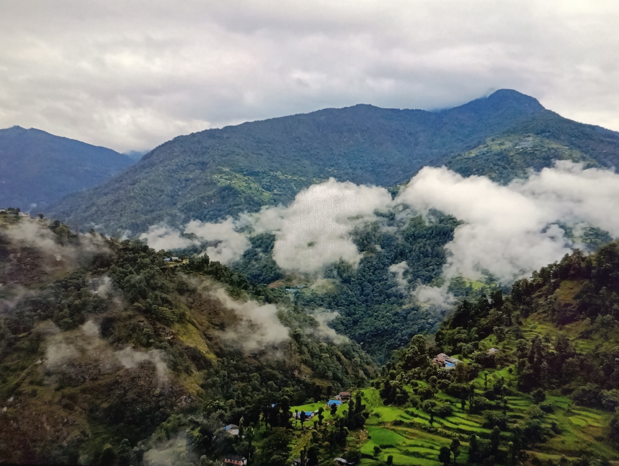 This is an aerial, high-angle, medium shot of a mountainous landscape with rolling green hills, dense forests, and scattered clouds, under a cloudy sky. In the foreground and midground, several small clusters of houses with blue roofs are visible, nestled amongst the trees and on terraced green fields. The clouds appear to be partially obscuring some of the higher peaks in the background. The scene conveys a sense of a rural, possibly remote, mountainous region. The location in the city of Dubăsari town council, Moldova is not discernable from the image itself.