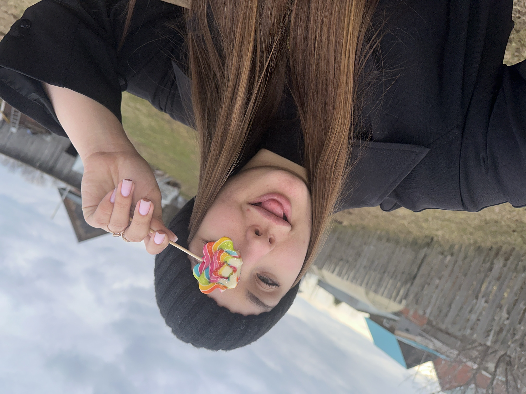 A young woman stands outdoors, holding a multicolored flower-shaped lollipop over her left eye. She wears a black beanie and a black coat. Her hair is long and brown. Her tongue is slightly visible, protruding from her mouth. She is wearing a ring on her left hand. The background shows a fence and buildings. Based on the landscape and architecture, the location is likely Ludorvay, Russia. The sky is overcast.