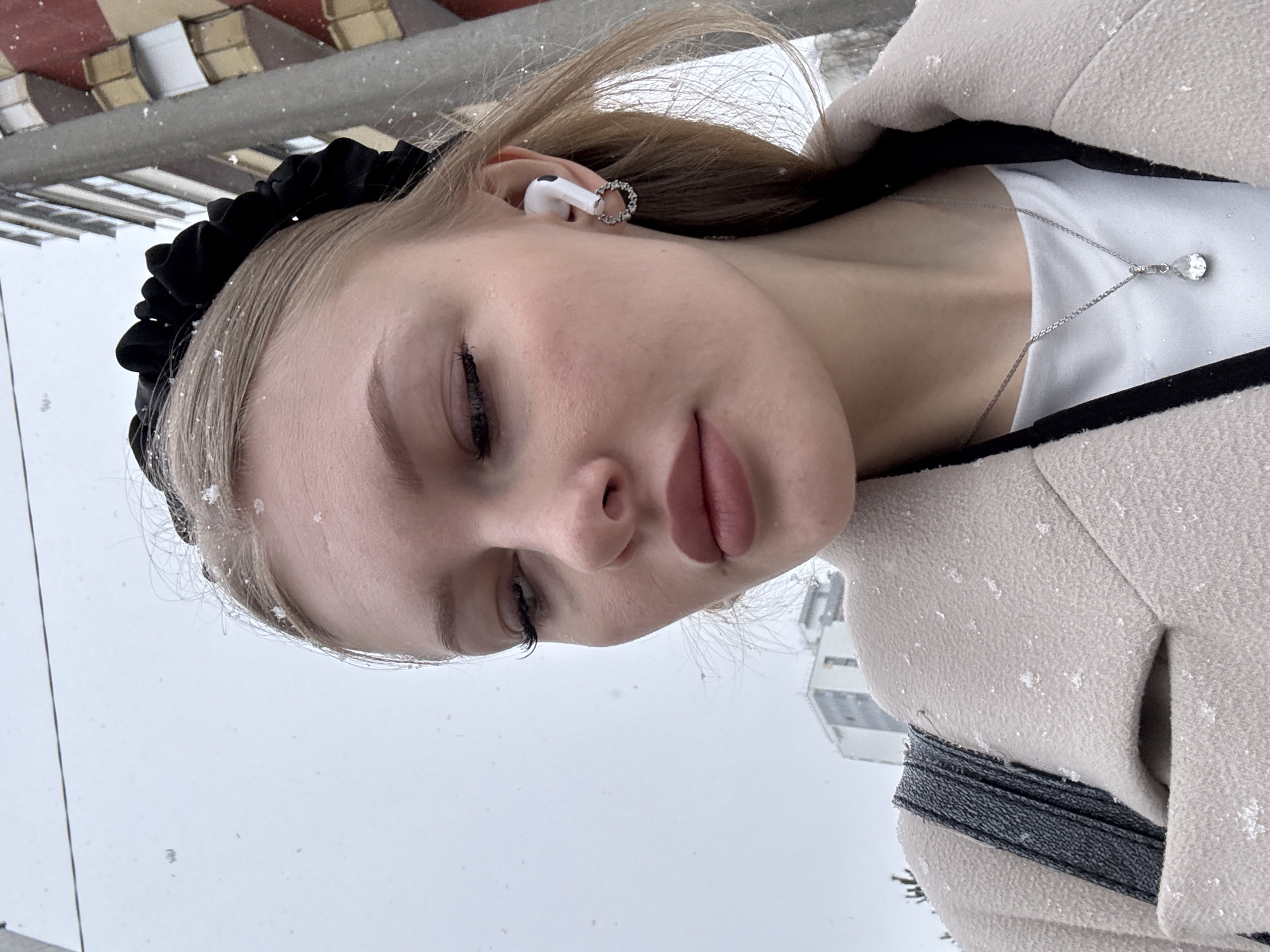 A close-up portrait, oriented on its side (rotated 90 degrees counter-clockwise), captures a young woman outdoors in Perm, Russia, during a light snowfall. Small white specks, indicative of snowflakes, are scattered across her light-colored hair, skin, and coat. She wears a black ruffled headband or scrunchie and has a serene or contemplative expression with her eyes mostly closed or looking downwards. Her lips are naturally parted. In her right ear, she wears a white wireless earbud and a silver-toned ear cuff. A silver-toned necklace with a clear or white tear-drop shaped pendant adorns her neck, resting on a white top visible beneath a light beige or cream-colored coat with a dark trim. A black strap, likely from a bag, is also visible. The background is soft-focused, showing an overcast sky and indistinct urban architecture, consistent with an outdoor winter scene. No text is visible in the image.