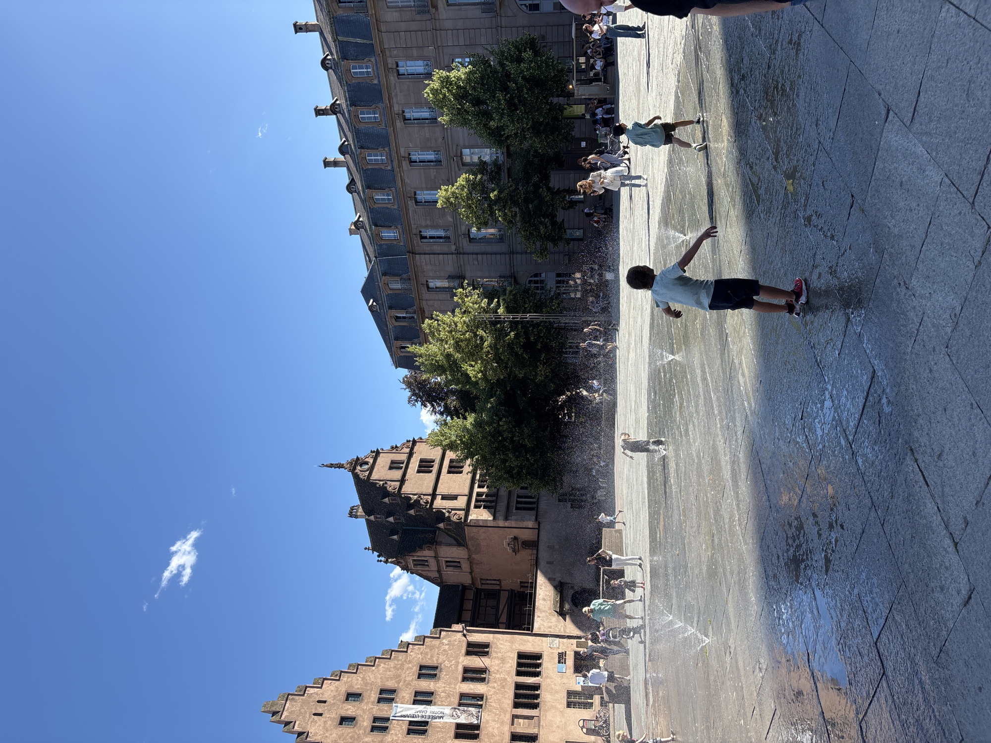 The image depicts a sunny day in Strasbourg, France, with a clear blue sky. The scene is set in a plaza with a water fountain feature, where several children are playing and getting wet. The ground is paved with large stone tiles, wet from the fountain spray, creating reflections of the surrounding buildings and trees.
In the background, a prominent building with a distinct stepped gable roof, characteristic of Alsatian architecture, stands tall. This building is likely part of the historic city center. To the right, a larger, multi-story building with many windows and a dark mansard roof lines the plaza. Lush green trees are scattered throughout the scene, adding to the pleasant outdoor atmosphere.
Several adults and children are present in the plaza, some watching the children play, others walking by. A banner is visible on the left side of the stepped gable building, possibly advertising an event or attraction. The overall impression is a lively public space enjoyed by locals and visitors alike on a summer day.