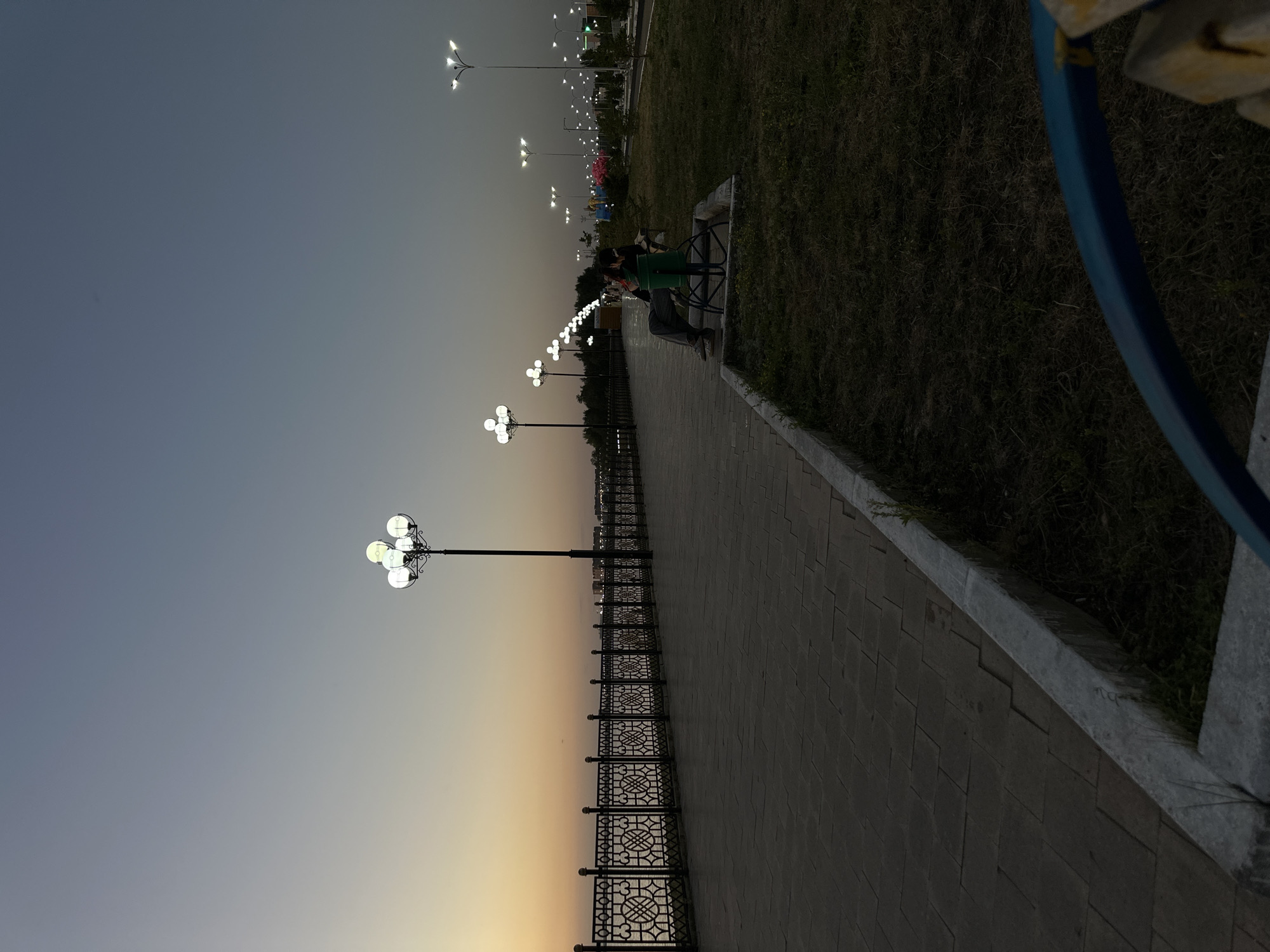 The image depicts a twilight scene along a waterfront promenade in Kokshetau, Kazakhstan. A paved walkway runs alongside a decorative metal fence overlooking a body of water. Several ornate lampposts with multiple lights illuminate the pathway, casting a warm glow. In the middle ground, a couple is seated on a bench in a grassy area. Beyond them, more lampposts line a distant road, and in the far background, the lights of the city are visible. The sky is a gradient of soft blues and oranges, indicative of sunset or sunrise.