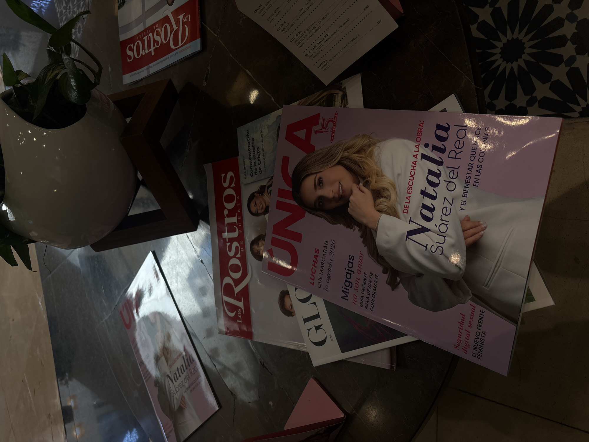 The image captures a collection of items scattered on a dark surface, likely a table, in Puebla, Mexico. A white ceramic pot containing a green plant sits on the left edge of the frame. Several magazines are present, with "Los Rostros" and "UNICA" being visible titles. The cover of one magazine features a blonde woman named "Natalia Suárez del Real". Also visible is a leaflet with the text "Está invitado Conmemoración de la muerte de Cristo Jueves 2 de abril de 2026". A partially visible menu shows a list of food items and prices. The background reveals a surface with a geometric tile pattern.