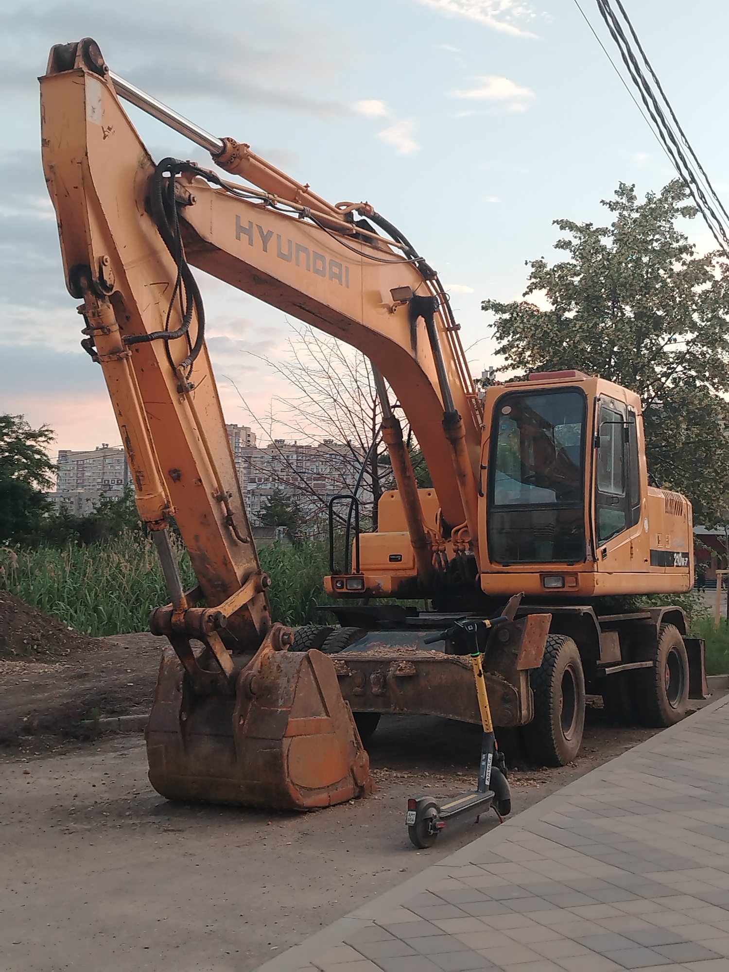 An orange Hyundai excavator is parked on a dirt and paved surface. The excavator has a large bucket and a visible "HYUNDAI" logo. In the background, there are tall buildings and trees. A yellow and black electric scooter is parked next to the excavator. The sky is partly cloudy with a hint of sunset. The scene appears to be an outdoor urban environment, likely a construction site or a street corner in Krasnodar, Russia.