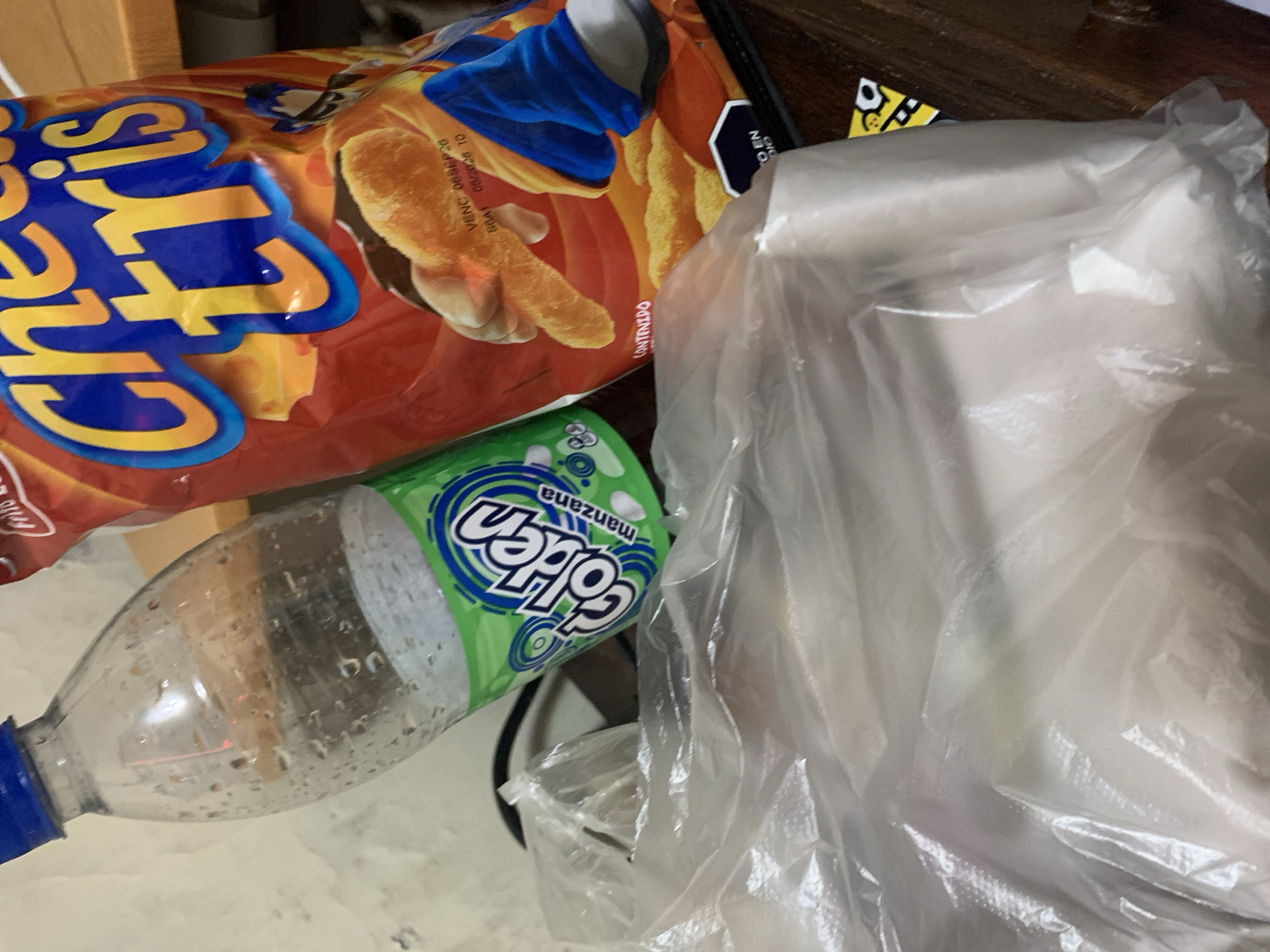 The image shows a collection of discarded items. A bag of "Cheetos" brand cheese-flavored snacks is partially visible. A plastic bottle, labeled "Golden Manzana" (Apple flavor), with a blue cap, is present. A semi-transparent plastic bag occupies a large portion of the frame. The items are resting on a light-colored, possibly tiled, surface. There are no people visible, nor any indications of specific actions or interactions. The environment is unclear, other than being indoors with artificial light. There is no contextual information suggesting a specific location within El Valle del Espíritu Santo, Venezuela.