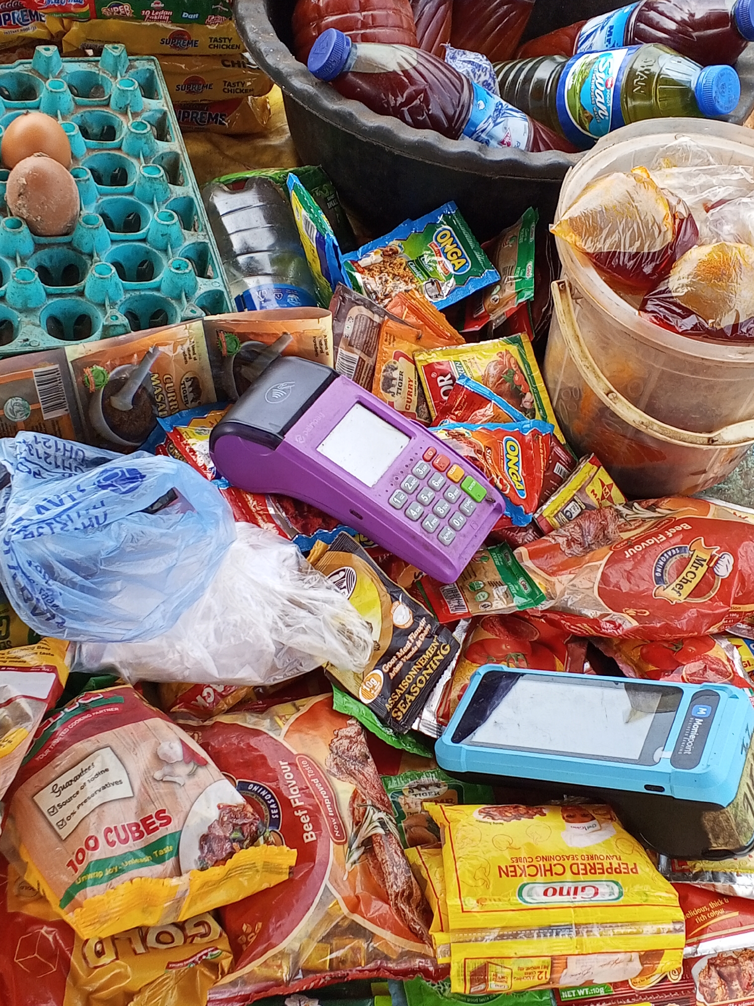 The image depicts a cluttered market stall in Gwong, Nigeria. There is no evidence of people, actions, or interactions; the stall appears unattended. Predominant objects include a blue egg carton containing two brown eggs, bottles of cooking oil, buckets containing what appears to be palm oil in plastic bags, and a variety of seasoning packets, including brands such as "Onga," "Mr. Chef," and "Gino." Several packages of "100 Cubes" bouillon cubes are also visible. A purple Palmpay point-of-sale device and a blue mobile phone with the "Moniepoint" logo are placed amidst the goods, suggesting a means of electronic payment. The environment is chaotic and crowded with various products.