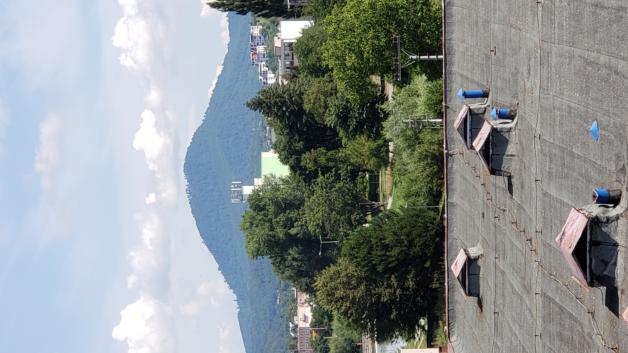 The image depicts a panoramic view of the city of Prešov, Slovakia, under a partly cloudy sky. In the foreground, a weathered, grey, flat roof is visible, featuring several square ventilation structures with red, pitched tops. These structures appear to be old and somewhat dilapidated. Two blue cylindrical bins and a blue conical object are also scattered on the roof.
Beyond the roof, a dense band of green trees occupies the mid-ground, obscuring much of the lower city. A river or body of water is partially visible to the left of the trees. In the background, several buildings of varying heights and architectural styles are seen, interspersed with more trees. A prominent white building with a green section stands out, topped with a structure that appears to be a telecommunications tower or antenna array. Further back, a large, verdant hill or mountain dominates the horizon, its peak crowned with what looks like another communication tower. The overall impression is of a city nestled within a natural landscape.