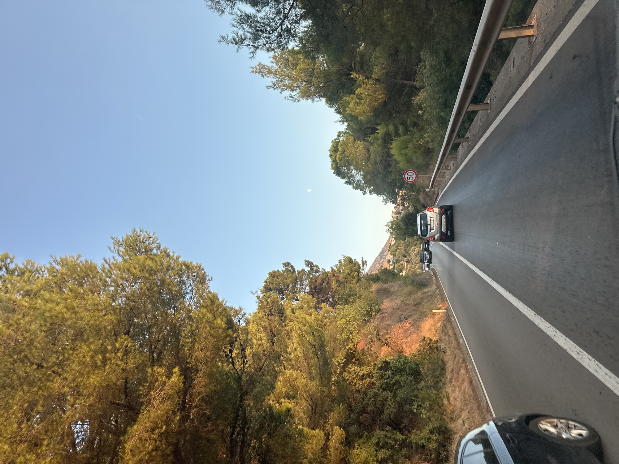 The image depicts a road in Sveti Stefan, Montenegro, with a clear blue sky above. Sunlight illuminates a steep, rocky hillside covered in dense green trees, with some sections catching a warm, golden glow. The road itself is paved, with a white line marking the center. Several cars are visible on the road. In the foreground, a black car is partially visible on the left side. Further down the road, a silver car is in the lane ahead, and other vehicles are further in the distance. A speed limit sign showing "50" is posted on the right side of the road, near a metal guardrail. The landscape suggests a mountainous or hilly terrain, with some buildings visible in the far distance on the right side. A faint crescent moon is visible in the sky.