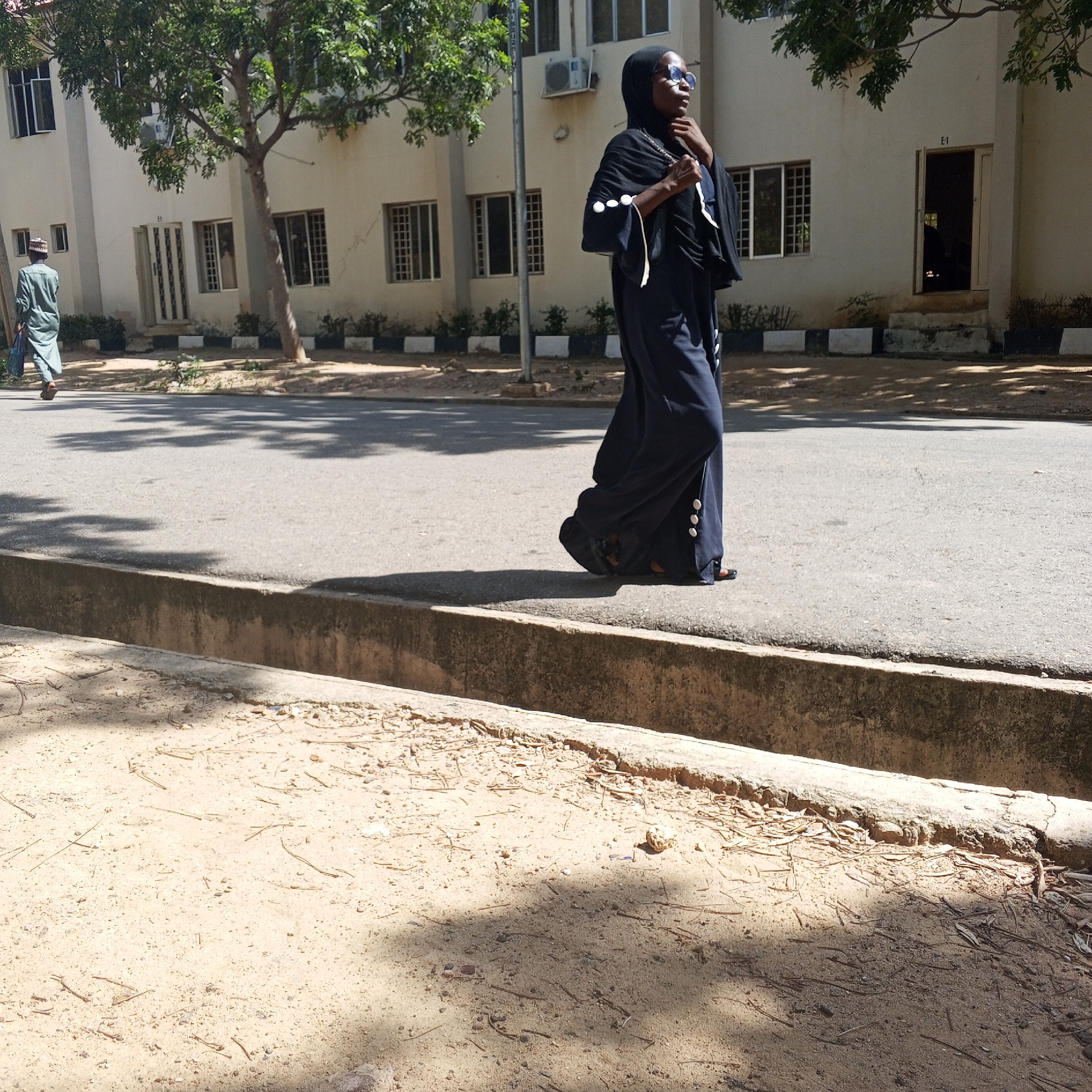 A wide shot captures a street scene in Arawa, Nigeria, on a sunny day. The foreground shows a paved road adjacent to a sandy embankment with scattered debris and pine needles. A concrete drain runs along the edge of the road. In the mid-ground, a person in a long, dark abaya with white embellishments on the sleeves walks on the street, facing right and looking to the left, wearing glasses and a hijab. Further back, another person in a light green traditional outfit walks away from the camera on the sidewalk, carrying a blue bag. Behind the sidewalk, a cream-colored building with multiple windows and an open doorway marked "E-1" is visible, partially shaded by a tree. The environment suggests an outdoor, urban setting with a mix of natural and man-made elements.