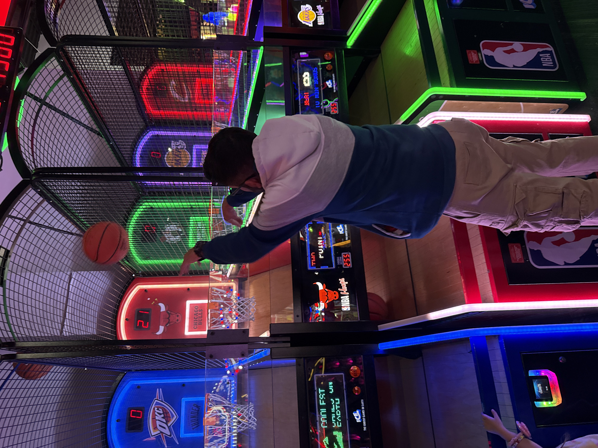 The image shows a person playing an NBA Hoops basketball arcade game. The game has multiple hoops with different team logos: Oklahoma City Thunder, Chicago Bulls, Los Angeles Lakers. The player is shooting a basketball towards the Chicago Bulls hoop, which currently has a score of 21. The score on the Oklahoma City Thunder hoop is 0. The person is wearing a blue and grey sweater and khaki pants. The game features illuminated LED lights and displays the high score as 255. The location of the game is the city of Columbus, United States of America.