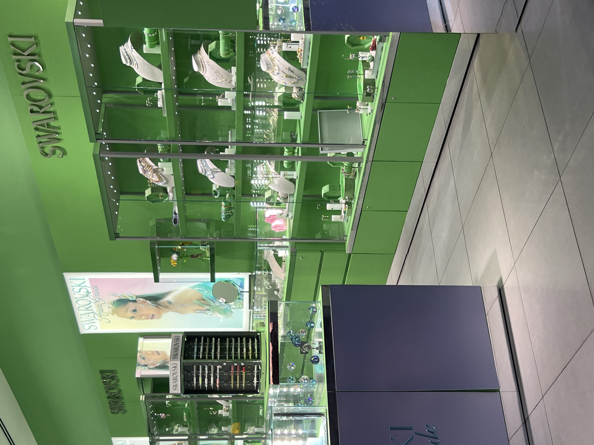 The image depicts the interior of a Swarovski retail store located in Kiryat Bialik, Israel. The environment is characterized by predominantly lime green walls and display unit bases, contrasting with a light grey tiled floor. Overhead, the "SWAROVSKI" brand name is prominently displayed in silver metallic lettering on the green wall.
The scene features multiple illuminated glass display cases arranged along the walls. These cases contain various jewelry items, including necklaces displayed on white and grey busts, earrings, bracelets, and rings, all set against green shelving.
A large, vertically oriented promotional banner is prominently featured, illuminated from behind, showcasing a female model identified as Ariana Grande, associated with "SWAROVSKI Ariana Grande" branding. A smaller, horizontal promotional image is also visible.
In the foreground, a purple-colored counter or display unit with a reflective metallic base is visible. On this counter, a clear glass rectangular container holds multiple faceted crystal spheres of varying colors, including blue, pink, and clear. To the left, a black rotating display stand holds multiple rows of what appear to be sunglasses or eyewear. No individuals are present in the depicted scene, and no actions or interactions are occurring.