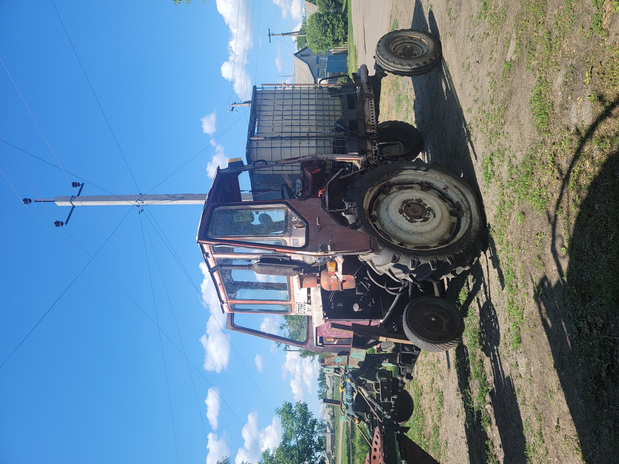 In Vovkivka, Ukraine, a weathered, red tractor is parked on a dirt and grass surface under a bright blue sky with scattered white clouds. The tractor, which appears to be old and in need of maintenance, is equipped with a large, square water tank mounted on its rear. A plow is attached to the front of the tractor. In the background, a concrete utility pole with electrical wires stands prominently. A few houses with pitched roofs and green trees are visible beyond the tractor, suggesting a rural or semi-rural setting. The lighting indicates it is daytime.