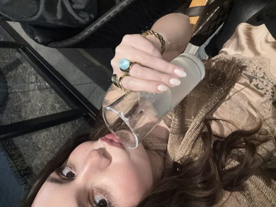 A woman holds a clear glass filled with a light-colored liquid to her lips. She has fair skin, dark hair, and wears rings on her fingers, including a large turquoise one. A gold bracelet adorns her wrist. She is dressed in a shimmering, gold-toned top. A portion of a black chair is visible in the background, with the lower left corner showing what appears to be a street sign.