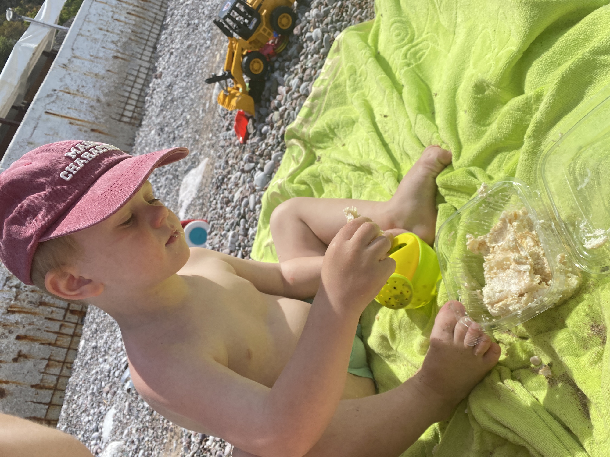 A young, fair-skinned child with blonde hair wearing a red baseball cap is sitting shirtless on a light green towel laid on a pebble beach. The beach appears to be in Rafailovići, Montenegro. The child holds a piece of food in their right hand, and a yellow watering can sits next to their left leg. A clear plastic container filled with food is placed near the child's feet on the towel. In the background, a yellow toy truck is positioned on the pebbles. A partial view of a concrete structure is on the far left of the scene.