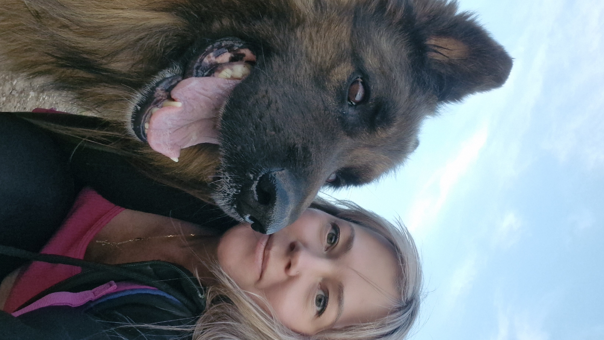 A low-angle, close-up photograph depicts a woman and a German Shepherd dog in an outdoor setting, likely daytime given the clear sky visible in the background. The woman, positioned at the bottom of the frame, faces the camera with a neutral expression. She has fair skin, light-colored hair, and is wearing a dark-colored jacket over a pink shirt. A thin necklace is visible around her neck. Above her, the dog's head dominates the frame. It has dark brown fur and its mouth is open, tongue lolling out, seemingly panting or smiling. The dog's nose is close to the woman's face, suggesting an affectionate interaction. The background consists of a light blue sky with some faint clouds. No specific urban or architectural features are visible to indicate the location is Otchor, Russia. The photograph captures a casual, candid moment between the woman and her dog.