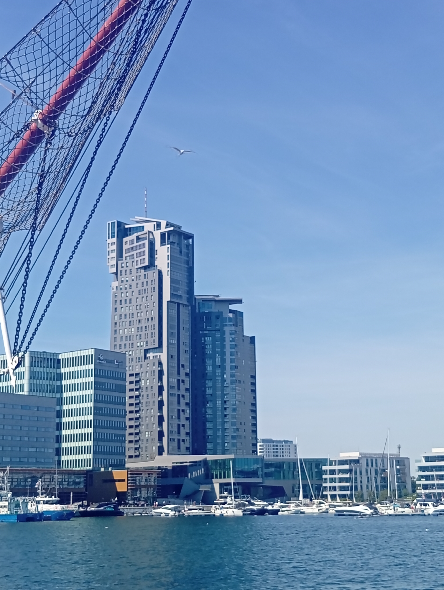The image depicts a waterside view of the city of Gdynia, Poland, with a focus on modern architecture and maritime elements. In the foreground, the rigging of a large ship is visible, with a red mast and black netting. The sea stretches out in front of the buildings.
Dominating the skyline are several tall, contemporary buildings. The most prominent is a multi-story skyscraper with a distinctive, stepped design, featuring numerous windows and contrasting grey and white facades. To its left, a slightly shorter, boxy building with a grid-like pattern of windows stands out. Other modern buildings, with glass facades and balconies, line the waterfront.
A marina is visible in the middle ground, filled with various sailboats and smaller motorboats docked in the clear blue water. The sky above is a bright, clear blue, with a single bird flying high in the distance, adding a sense of scale and life to the scene. The overall impression is of a vibrant, modern urban environment integrated with its maritime setting.
