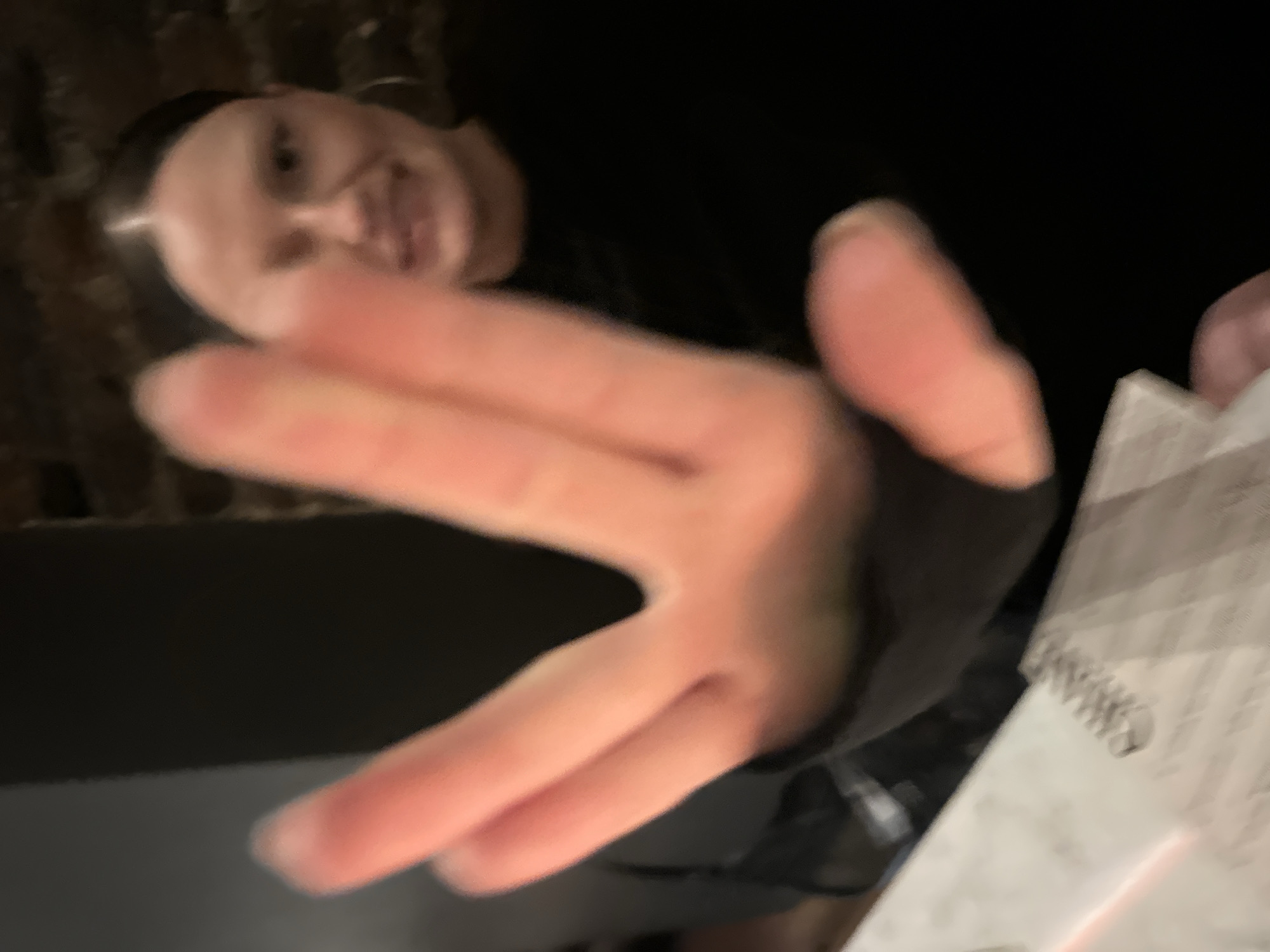 The scene depicts an indoor setting, consistent with a bar or restaurant, located within the city of London, United Kingdom, although the precise establishment is not discernible from the visible details. In the immediate foreground, a light-skinned human hand is sharply in focus, performing a "Vulcan salute" gesture with fingers separated between the middle and ring finger, and the thumb extended. Behind the hand, an adult female subject is visible, significantly out of focus, looking towards the camera with a discernible smile. She has dark hair with a center part, wears a dark long-sleeved garment, and is accessorized with hoop earrings. To the lower-left, a white document, appearing to be a menu or drink list, rests on a surface, with the word "CHAMPAGNE" partially legible in black text. The background features a dark, textured brick wall and a plain dark vertical surface, indicating a dimly lit environment with artificial light sources. The image exhibits a shallow depth of field, emphasizing the foreground hand.